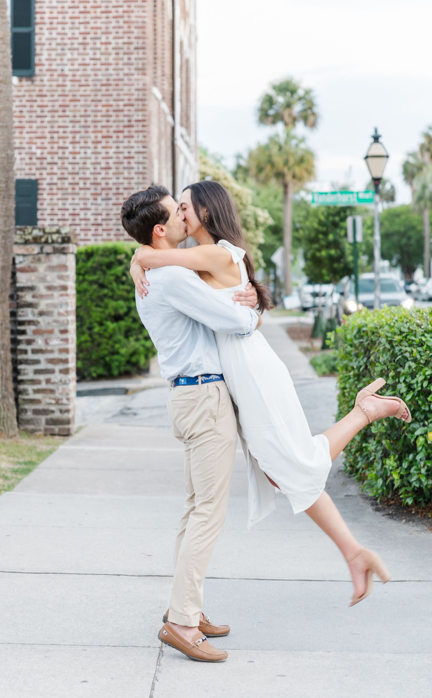 Man lifts woman for a kiss on a Charleston sidewalk, she wears a white dress and heels, brick building in background.