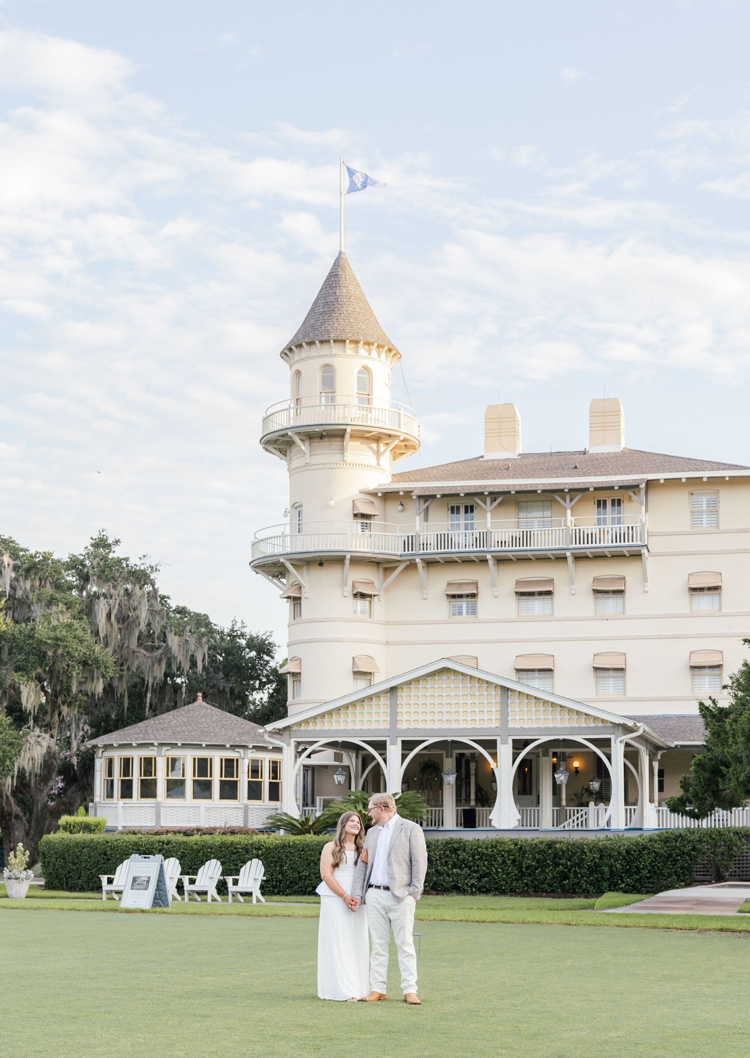 Couple poses in front of historic Jekyll Island Club Resort with turret and wrap-around porch on manicured lawn.