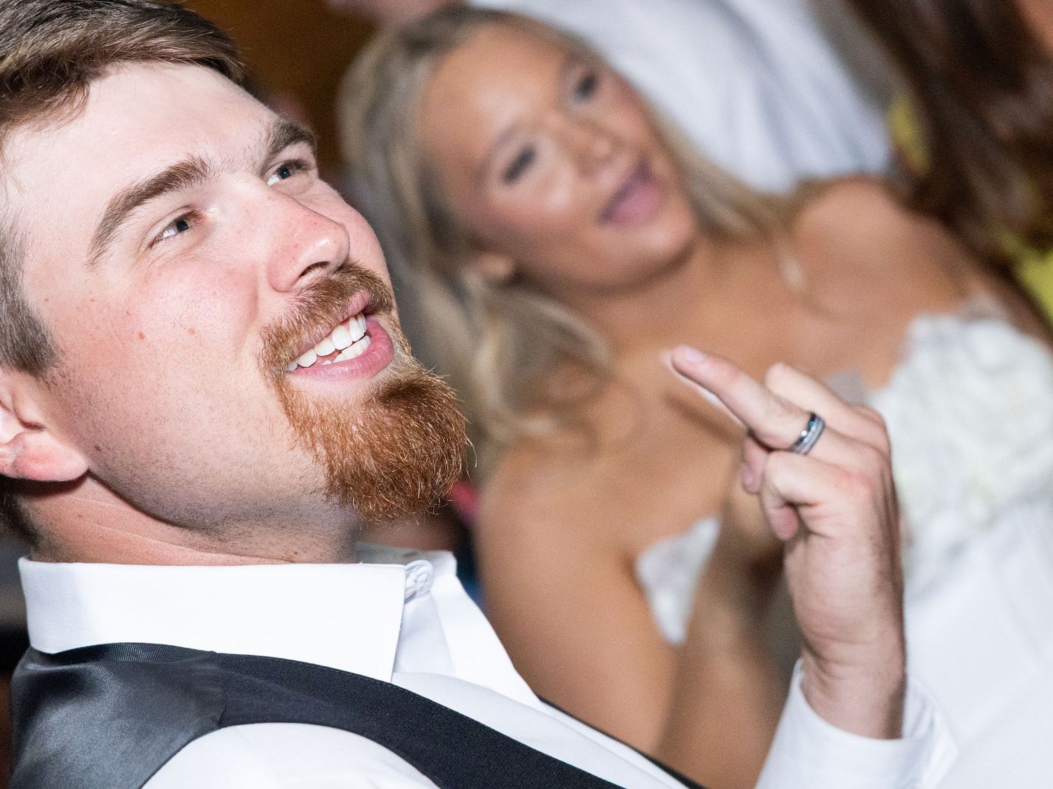 Smiling groom in black vest looks up while bride in white gown gestures playfully behind him.