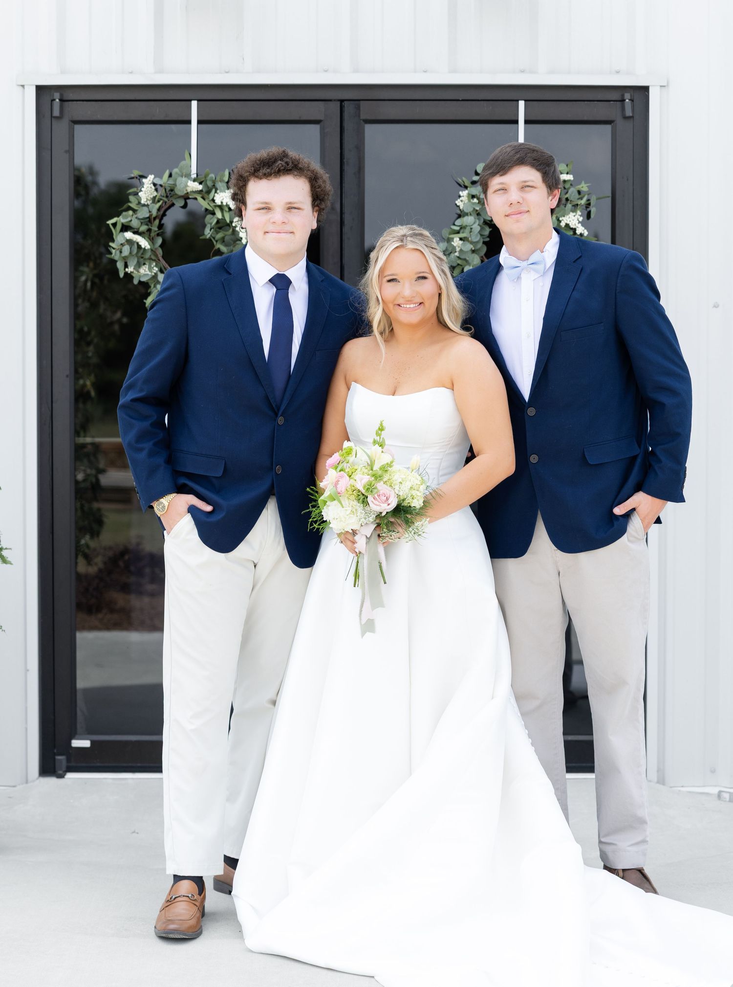 Smiling bride in white strapless gown holds bouquet flanked by two groomsmen in navy blazers outdoors.