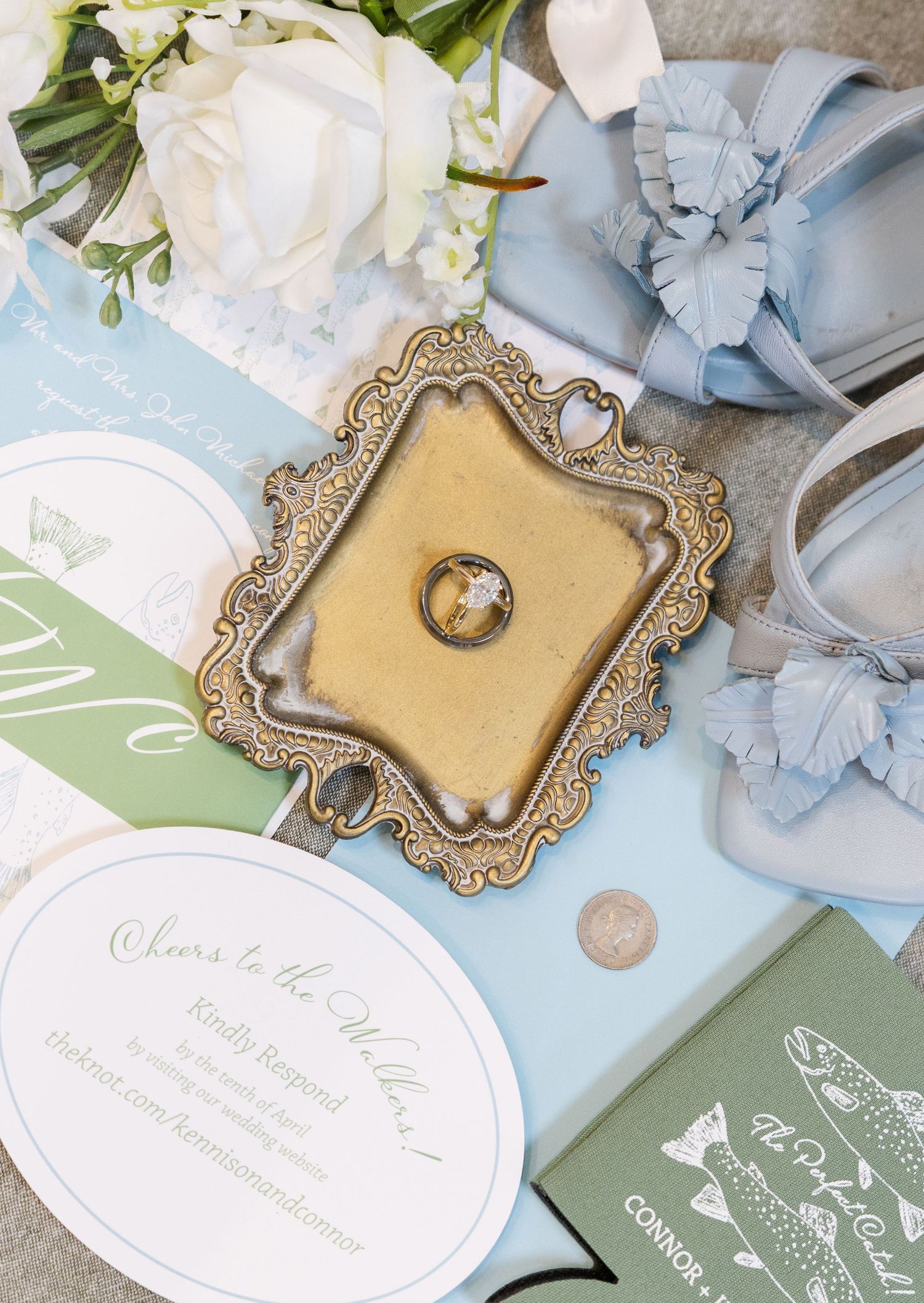 Elegant wedding flat lay with gold ornate frame, rings, blue heels, white flowers, and pastel green and blue stationery.