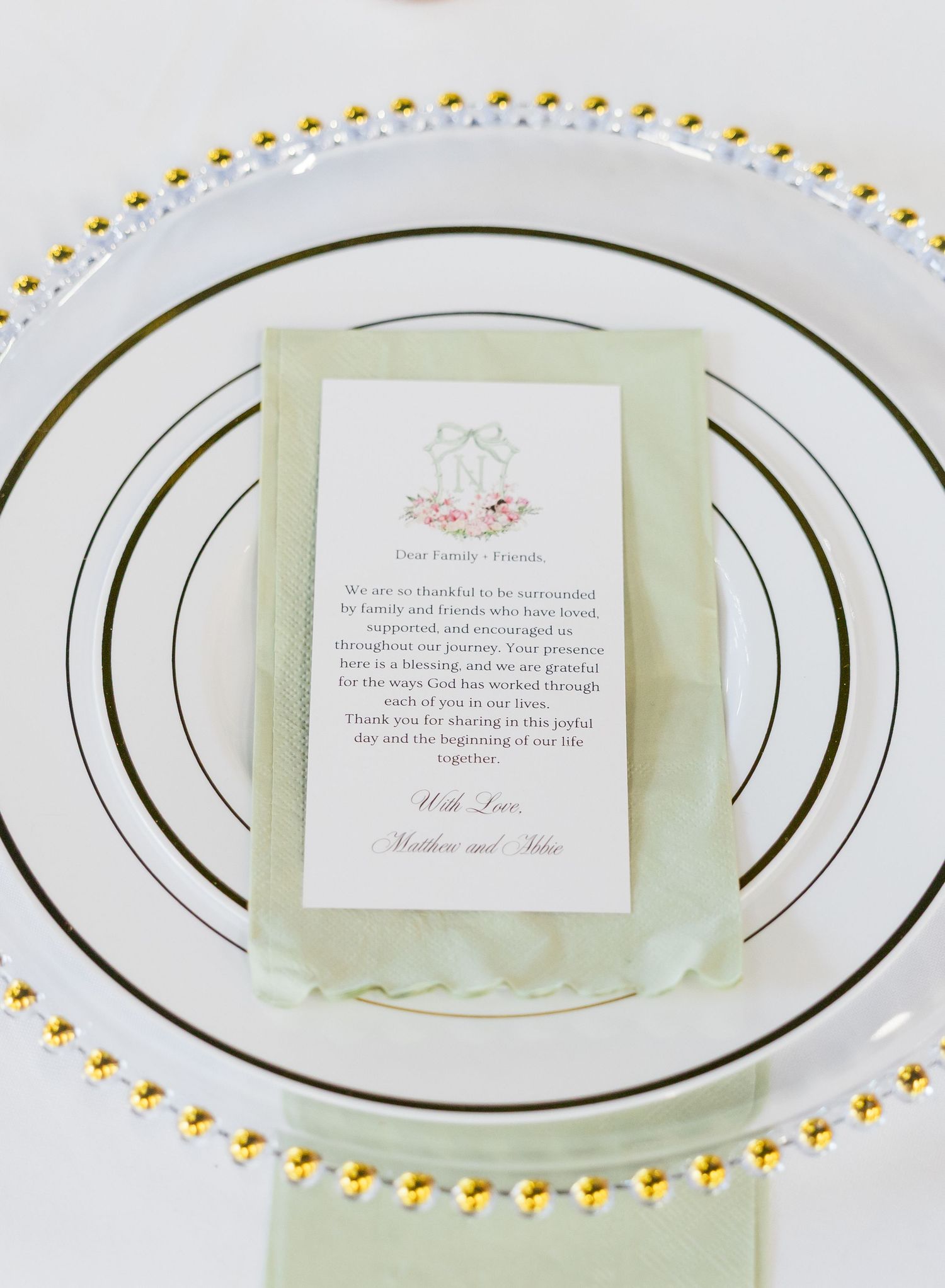 Elegant wedding place setting with gold beaded charger plate, sage green napkin, and personalized letter to guests.