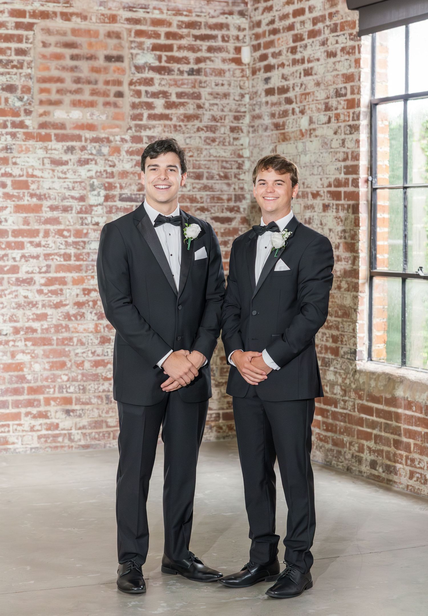 Two groomsmen in black tuxedos with boutonnieres standing before exposed brick wall.