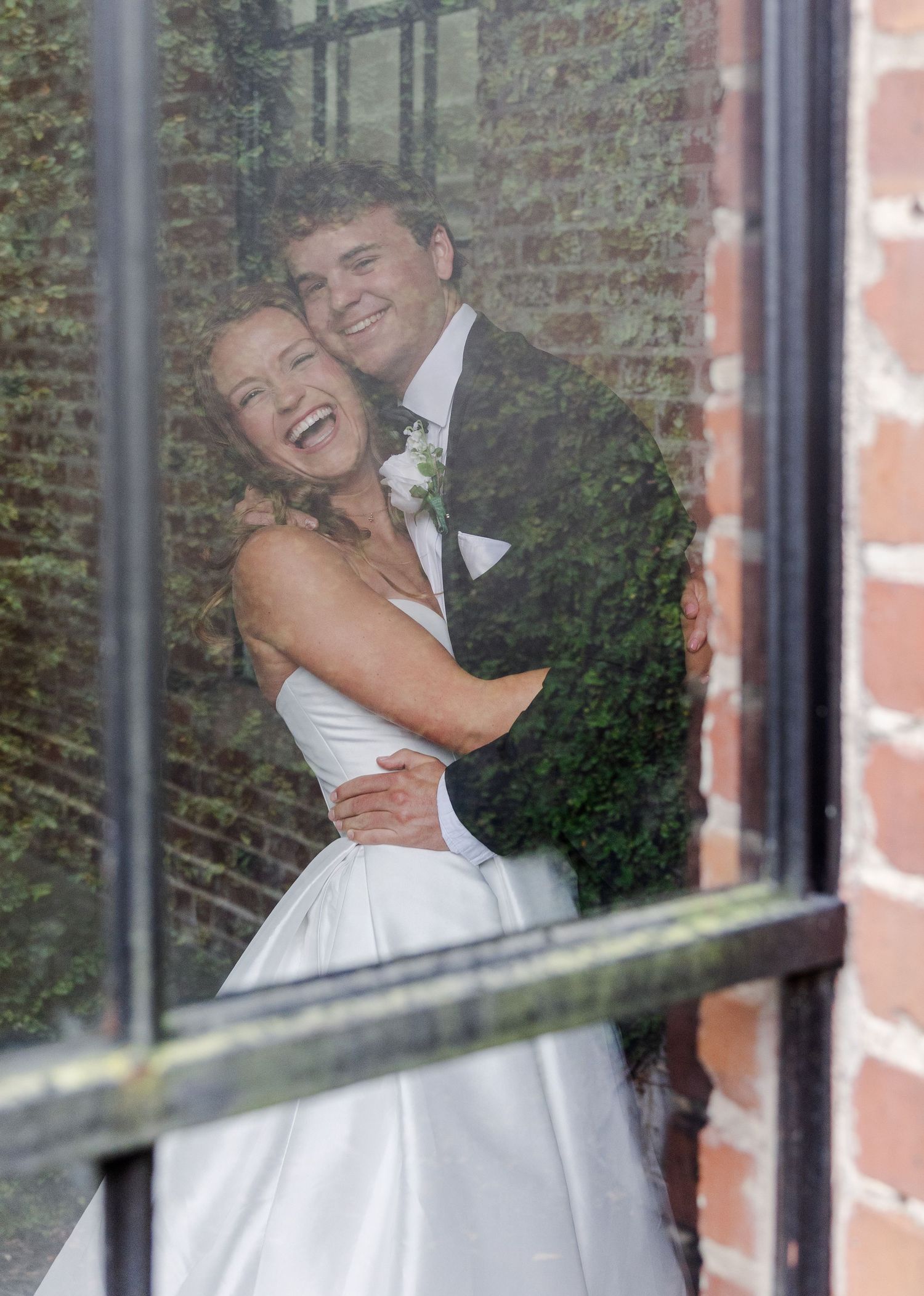 Joyful bride and groom embracing and laughing, captured through a window reflection.