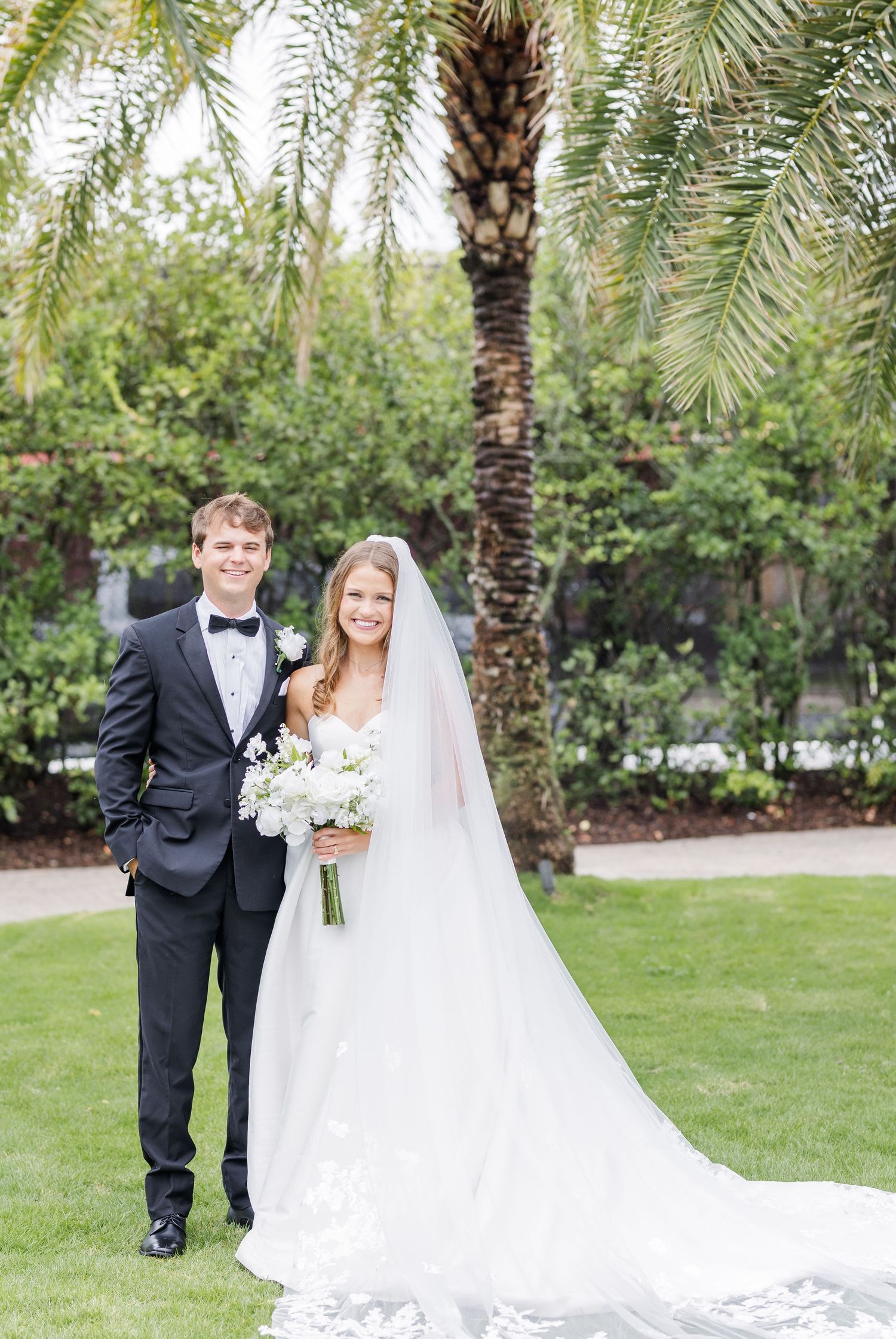 Bride in white ballgown with veil and groom in black tuxedo posing under palm tree outdoors.