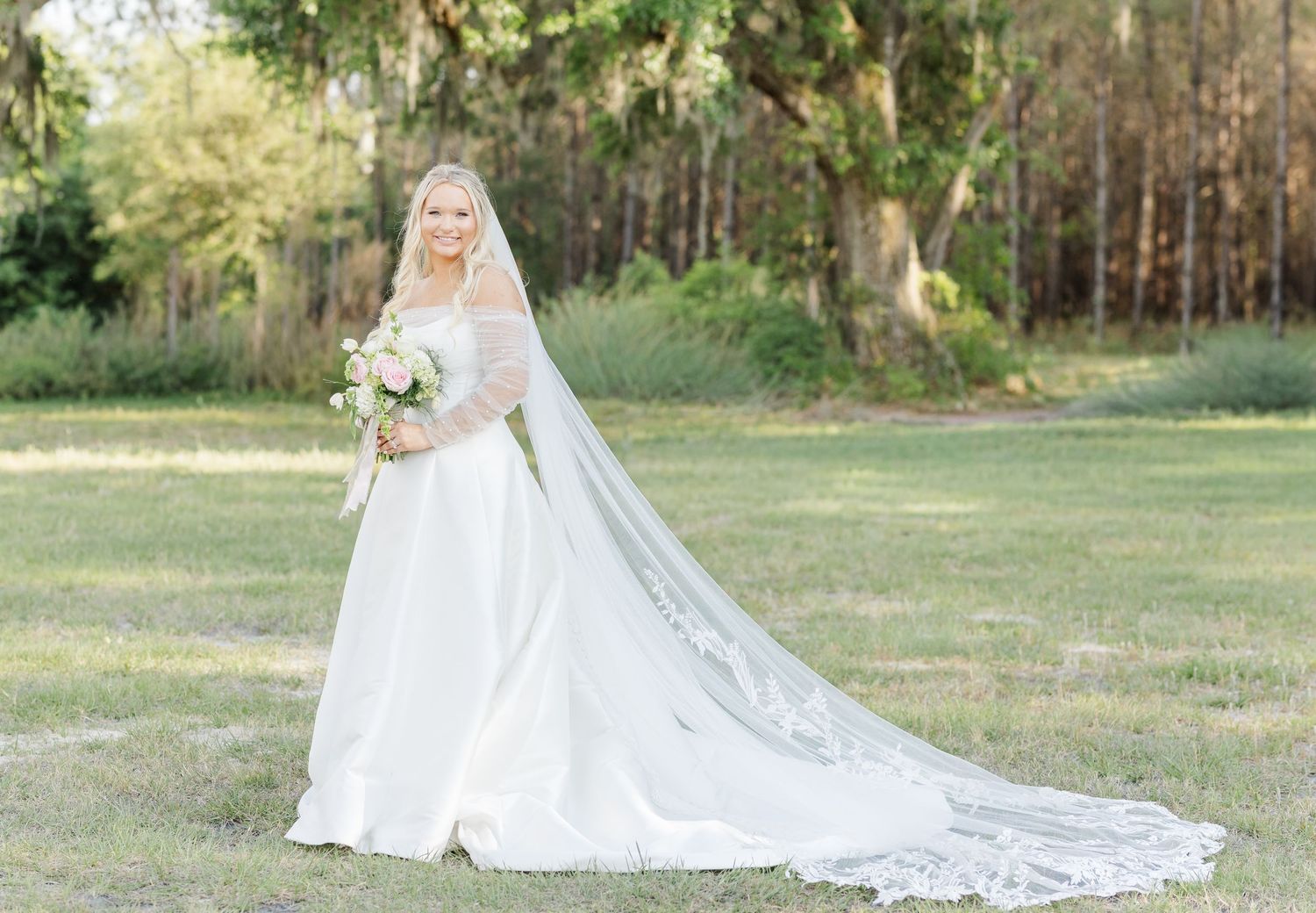 Bride in a long white gown with cathedral veil holding a bouquet in a green outdoor setting.