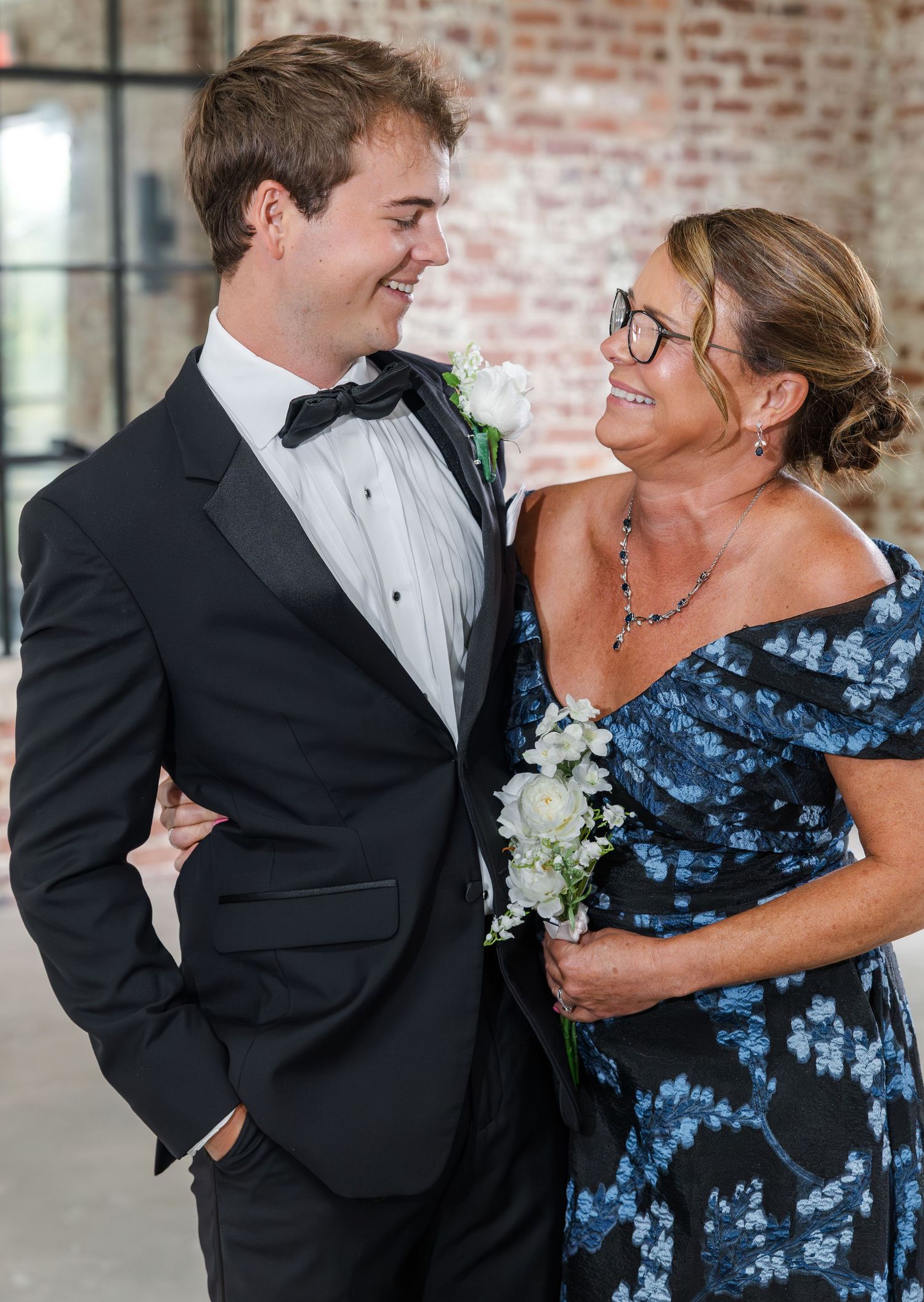 Groom in tuxedo shares a smile with mother in floral dress at wedding.