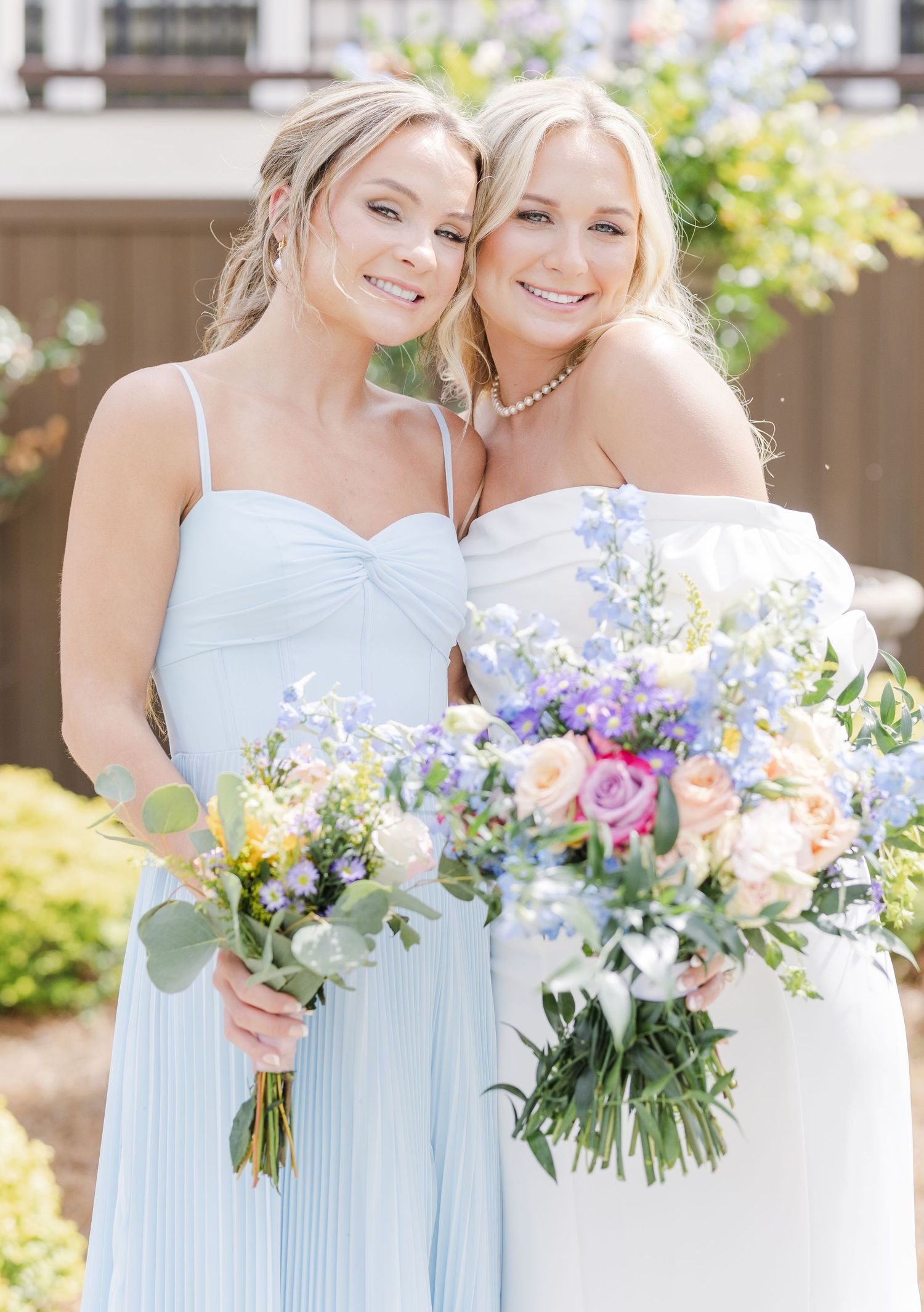 Bride in white strapless gown and bridesmaid in light blue dress holding colorful spring bouquets outdoors.