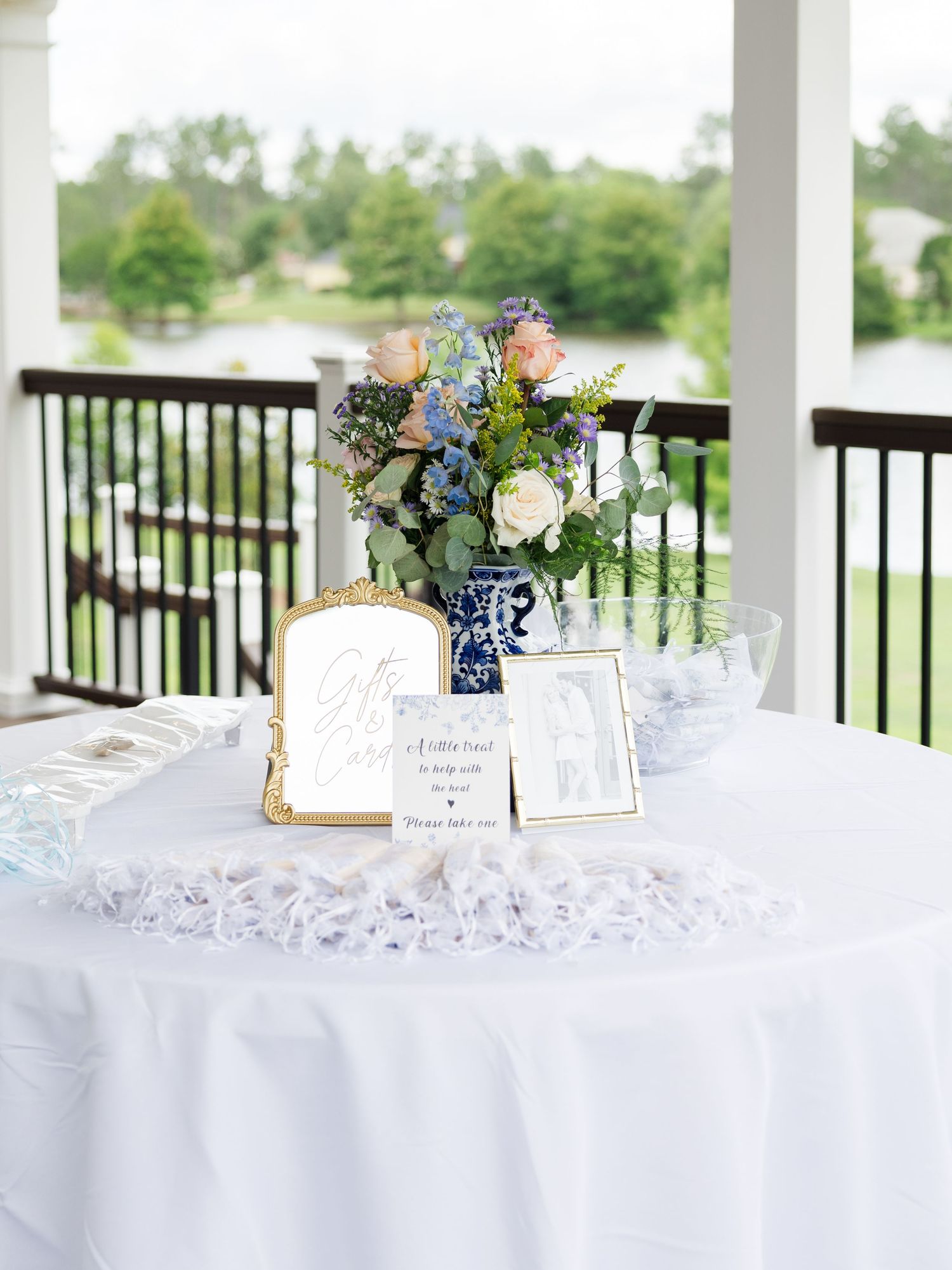 White outdoor wedding gift table with blue floral arrangement, framed photos, and signage on porch.