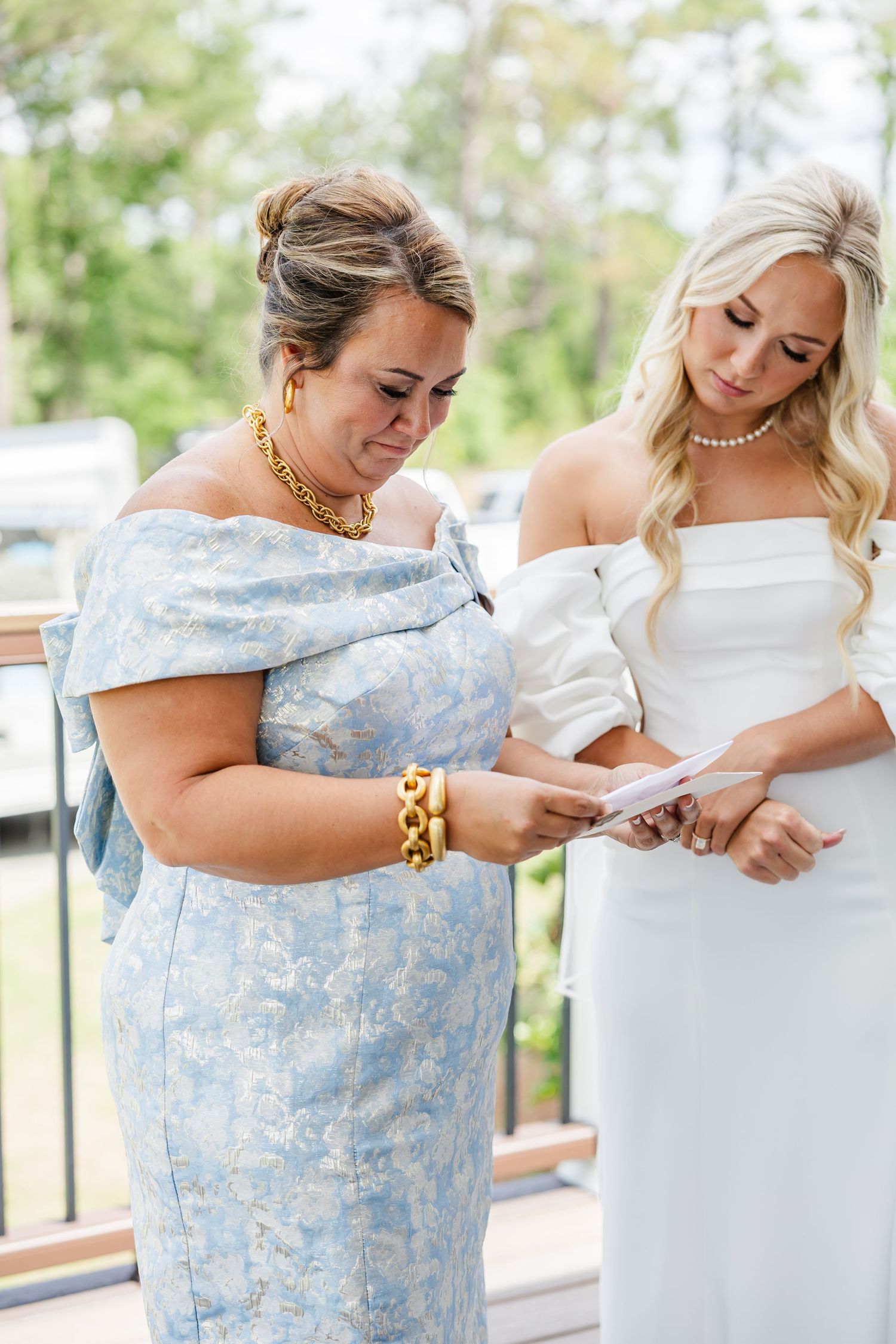 Bride in white gown reads note with emotional mother in blue floral dress on wedding day.