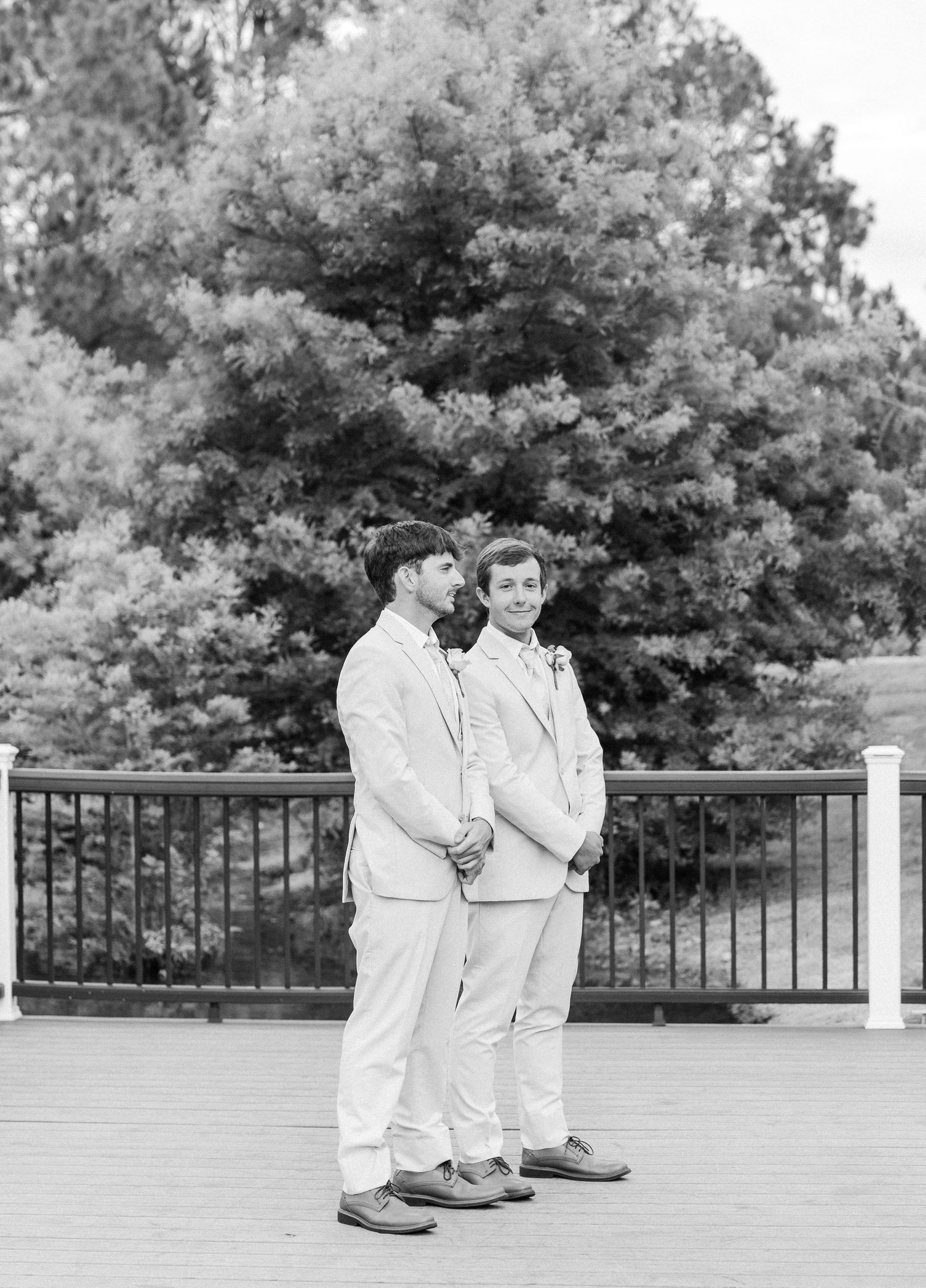 Two groomsmen in light suits stand together on a deck in a black and white wedding photo.