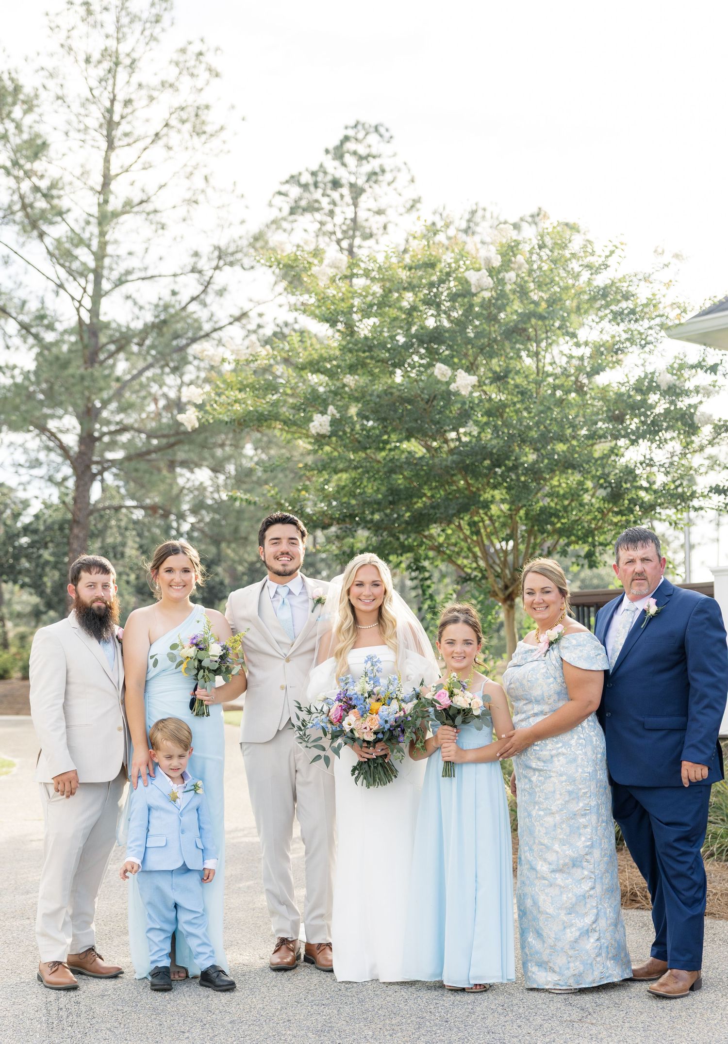 Wedding party group photo with bride, groom, and family members in coordinating blue and beige attire outdoors.