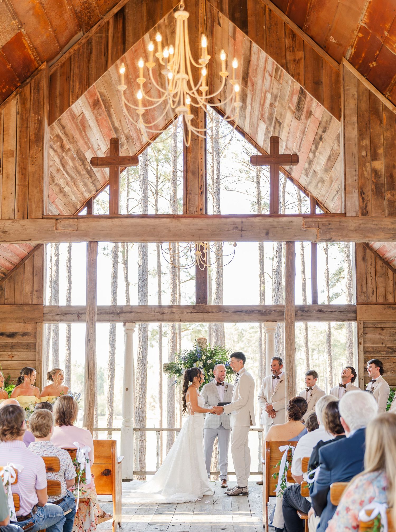 Bride and groom exchange vows in rustic wooden chapel with crosses and chandelier before seated guests.