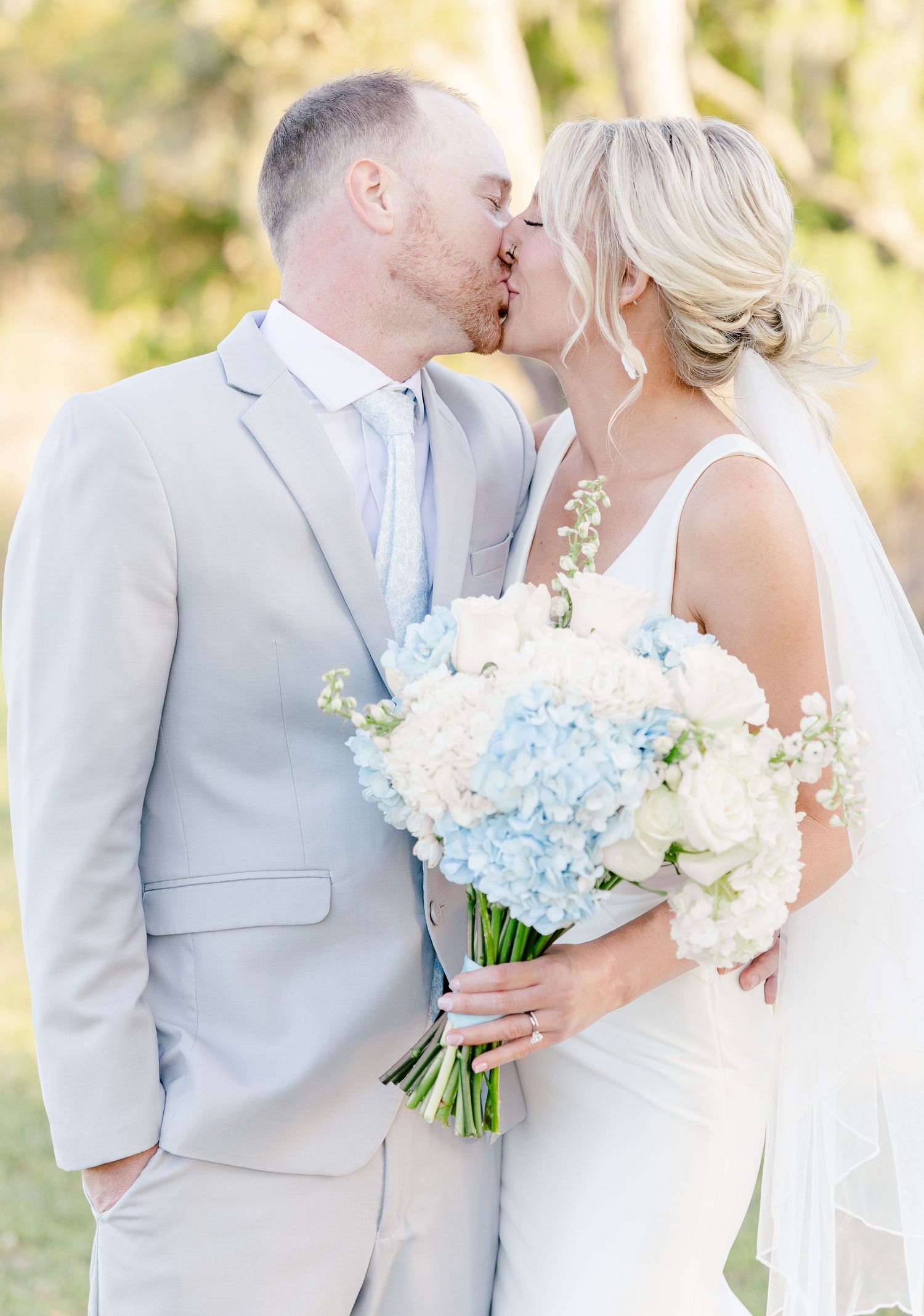 Bride and groom kissing with blue and white hydrangea bouquet at outdoor wedding.