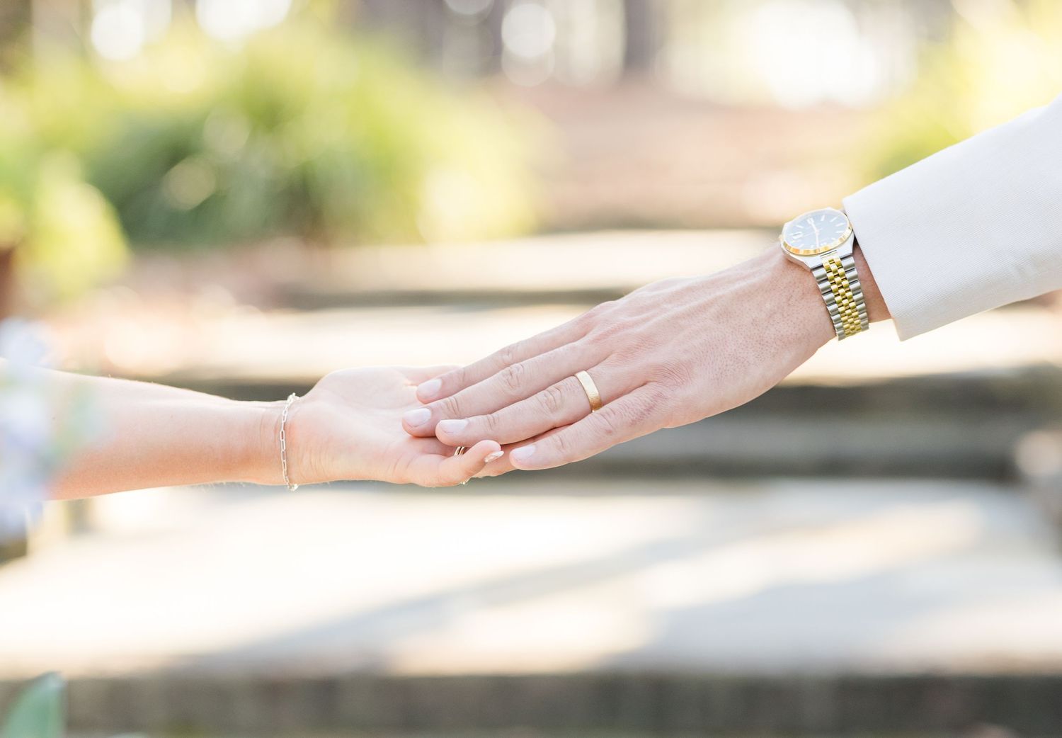 Close-up of newlywed couple holding hands, groom wearing wedding band and gold watch during outdoor wedding ceremony.