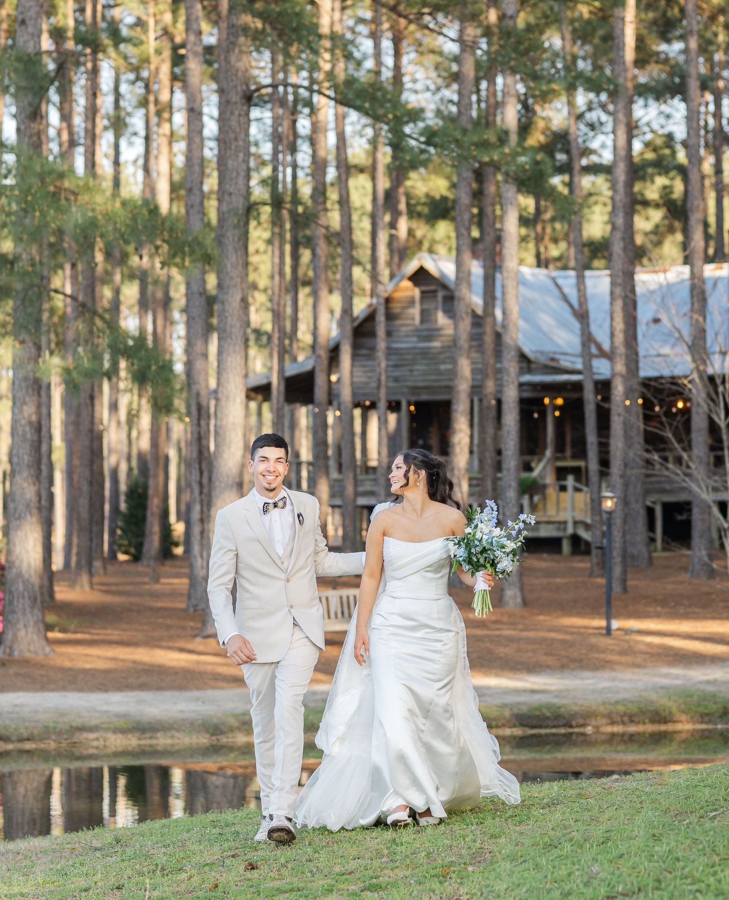 Bride and groom walk hand in hand near a scenic pond with a rustic log cabin venue amid tall pine trees.