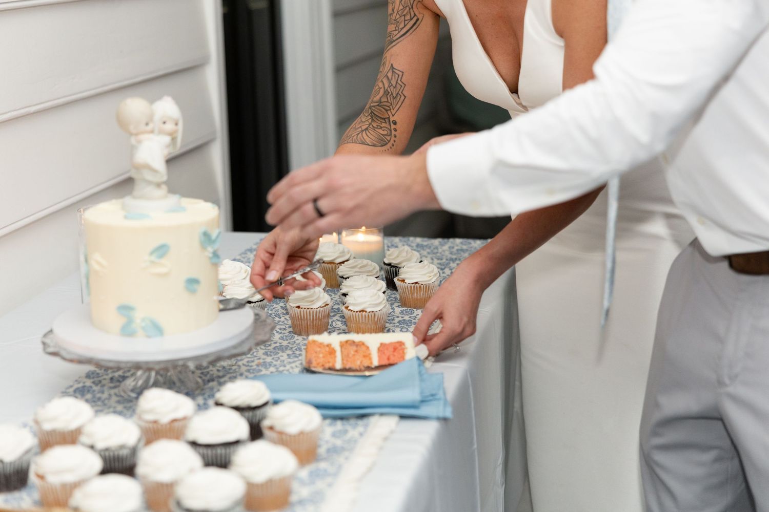 Bride and groom cut wedding cake surrounded by cupcakes at outdoor reception table.