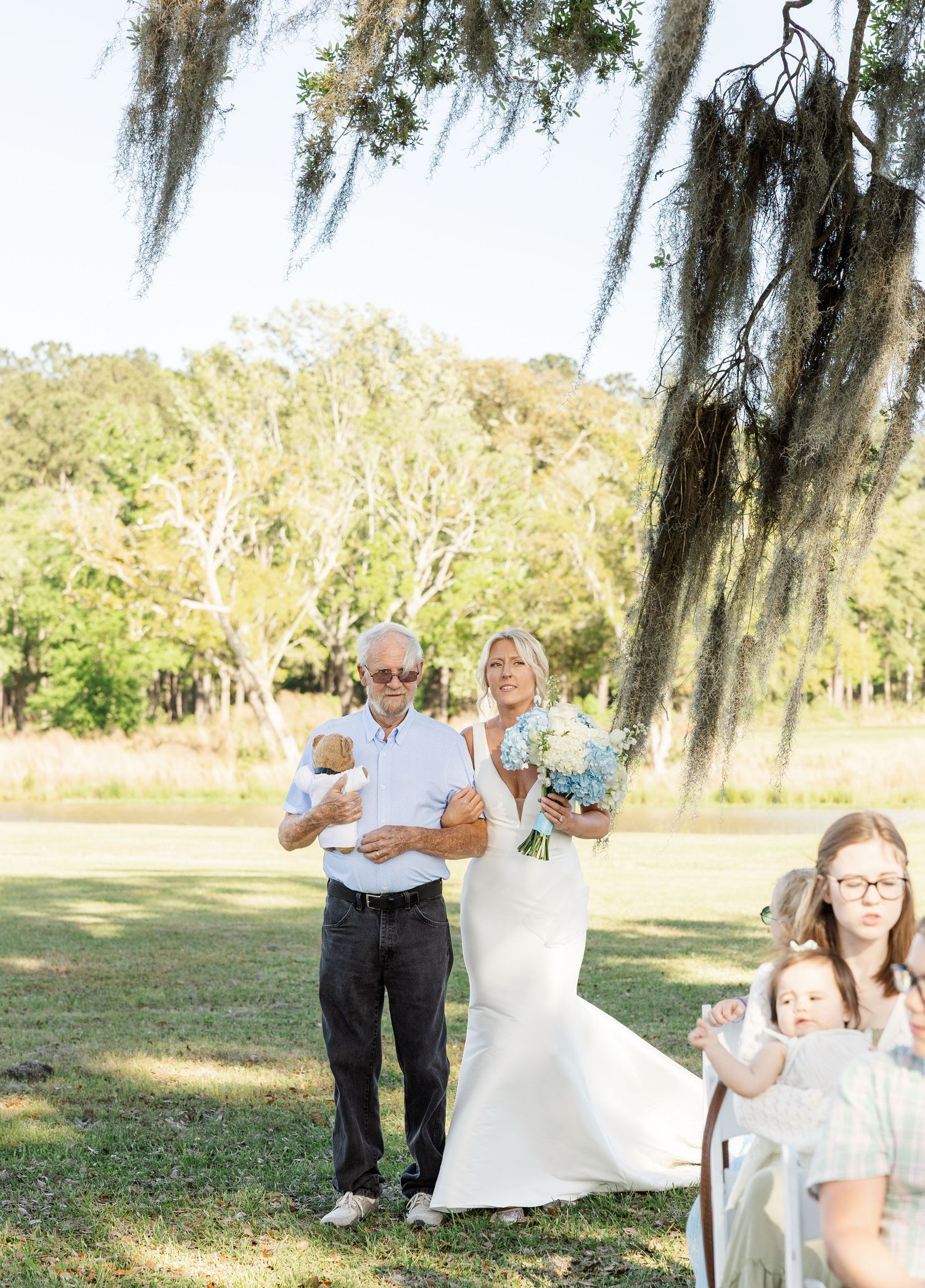 Bride in white gown holding blue bouquet walks down aisle with father under Spanish moss oak tree.