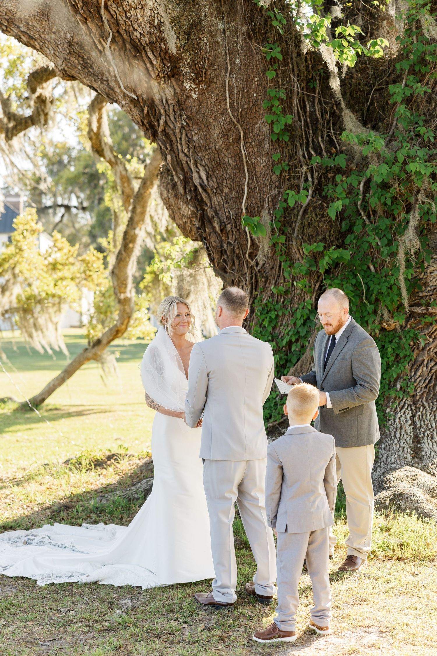 Bride and groom exchange vows under massive oak tree with young boy and officiant at intimate outdoor wedding.