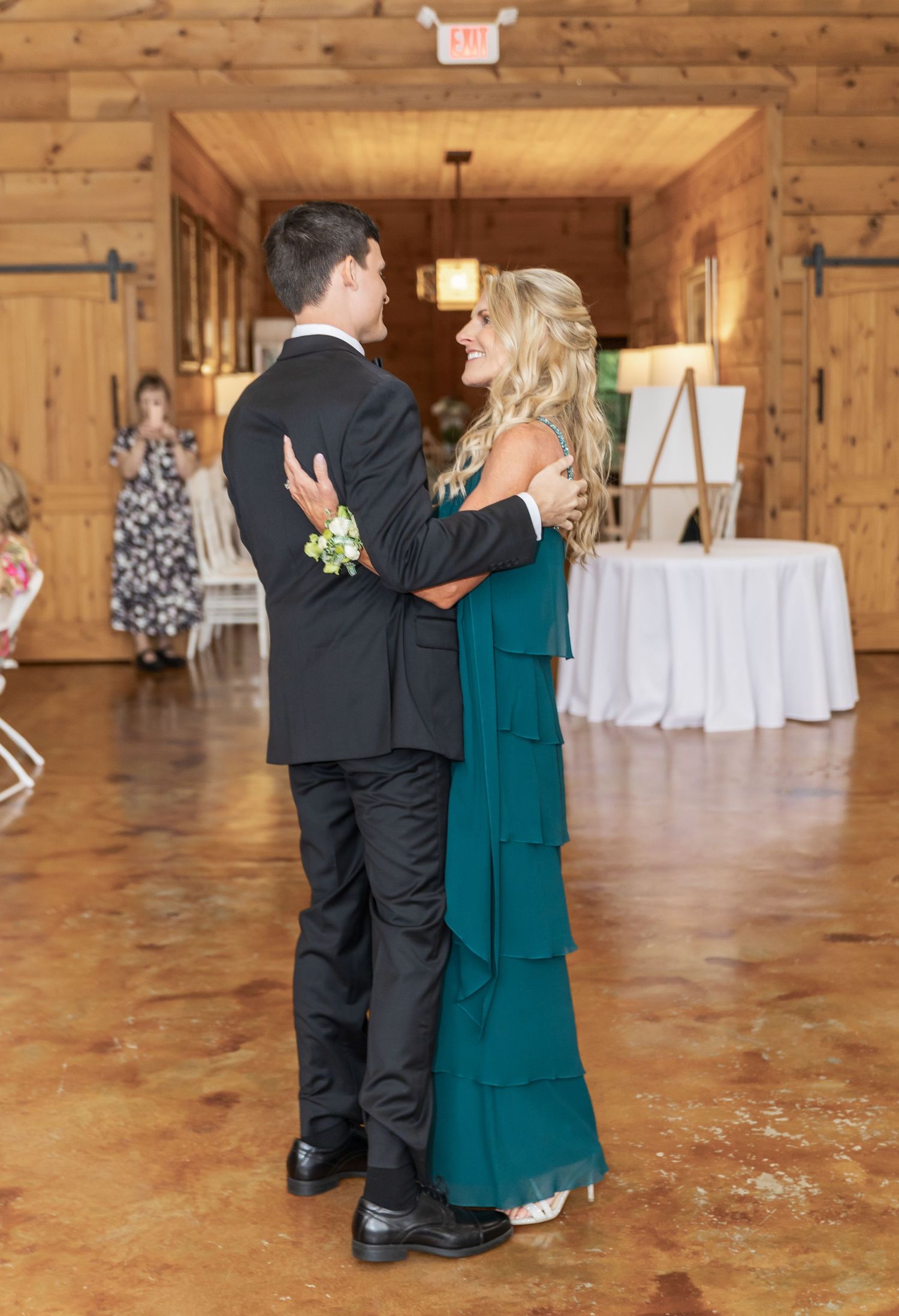Wedding guests dancing on wooden floor in rustic barn venue with white tables in background.