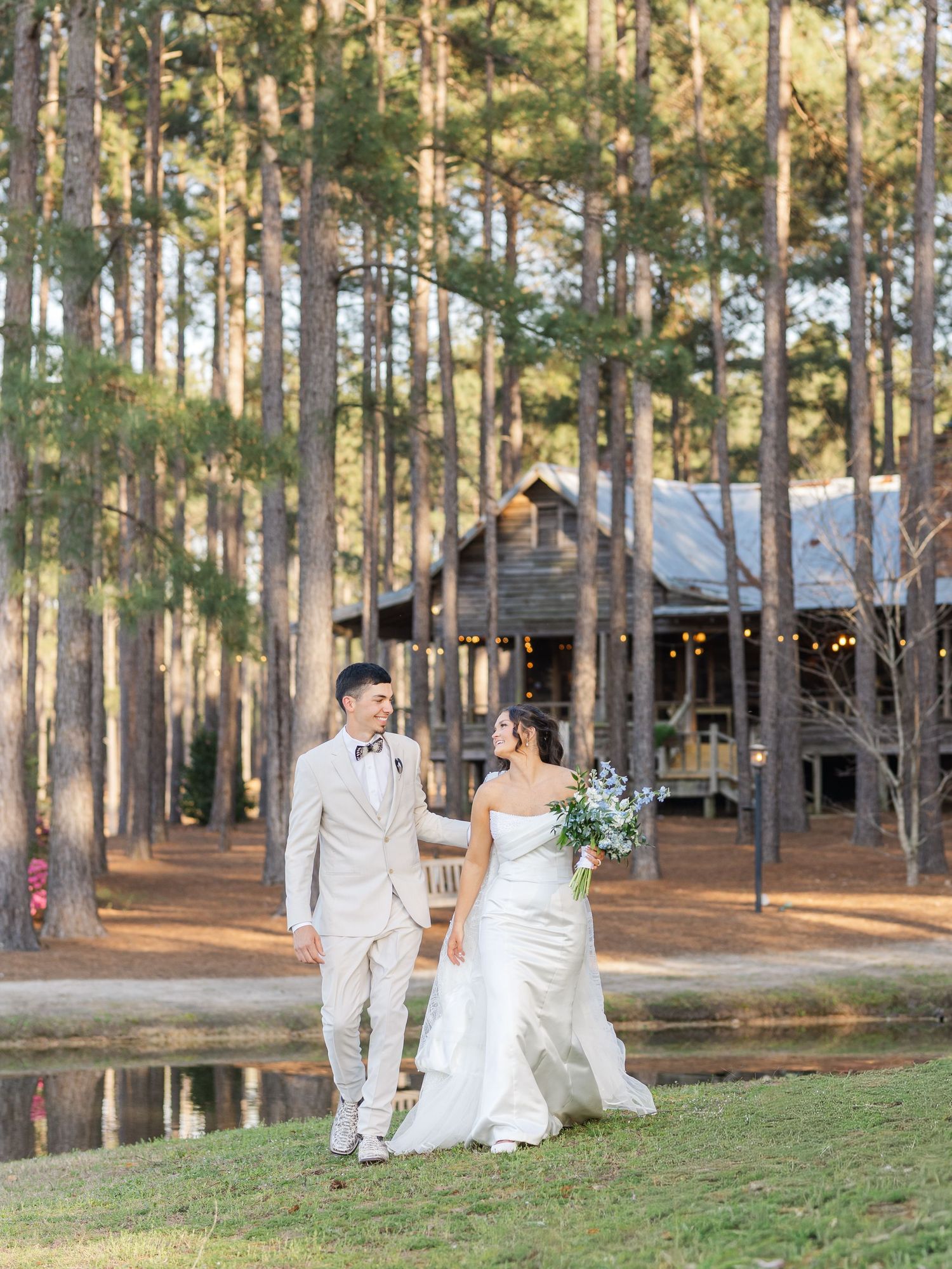 A couple in wedding attire walks hand in hand through a serene pine forest setting at sunset.