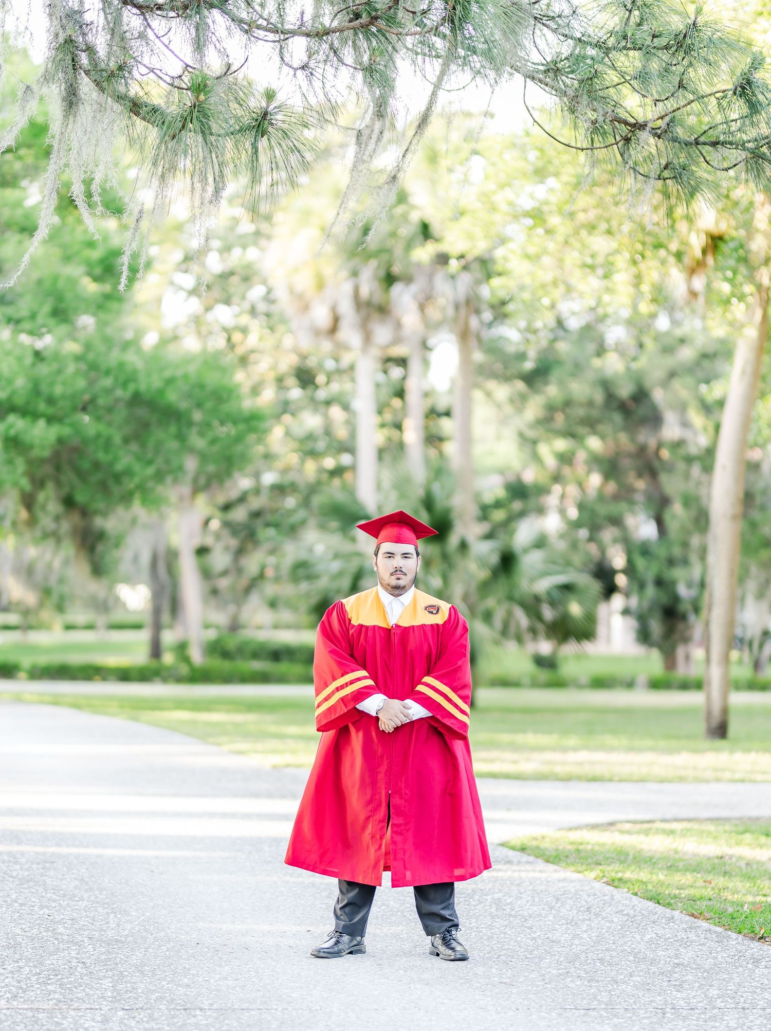 Graduate in bright red cap and gown stands on a tree-lined pathway during outdoor graduation photo session.