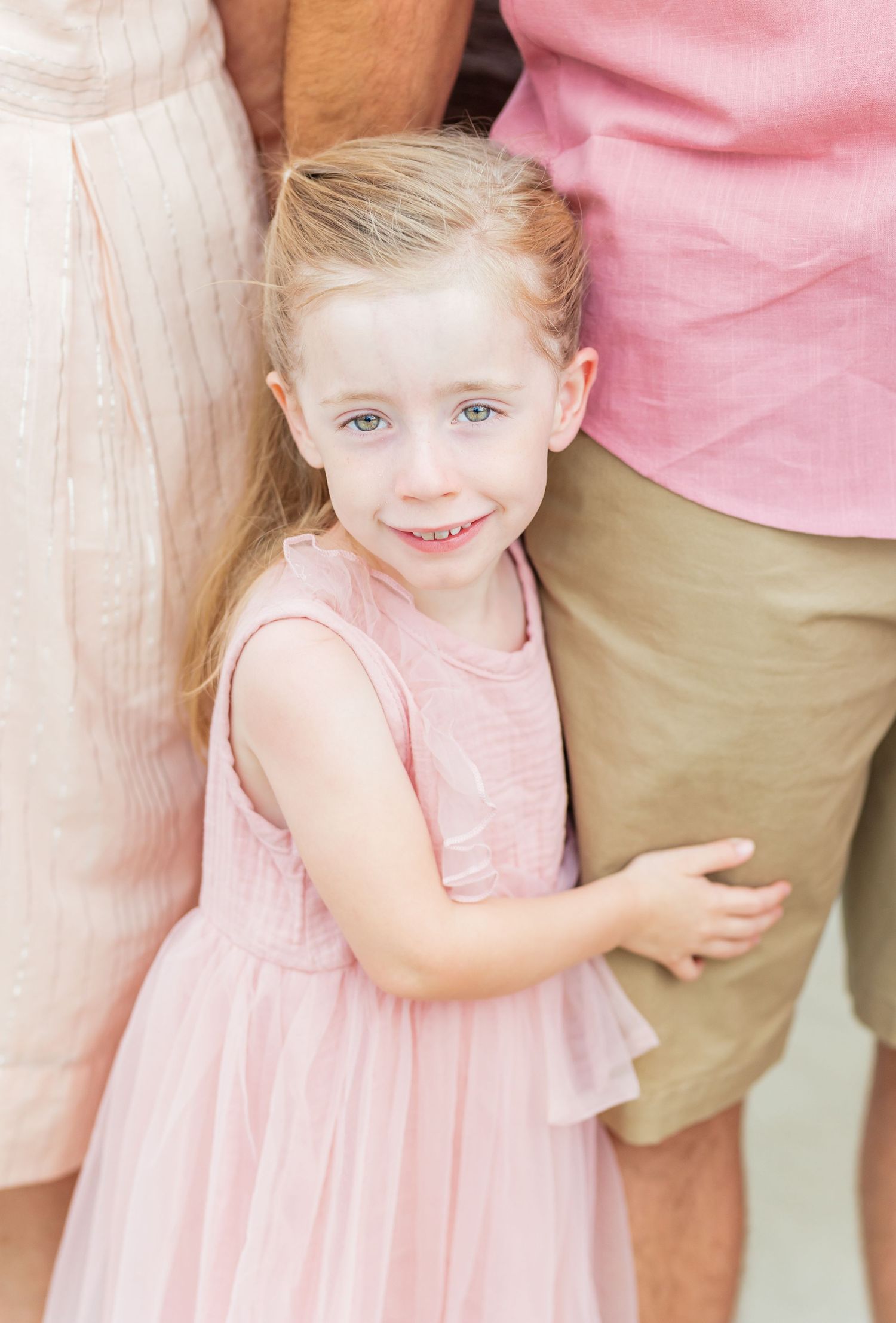 A small child in a pink sleeveless dress stands close to others wearing white and pink clothing.