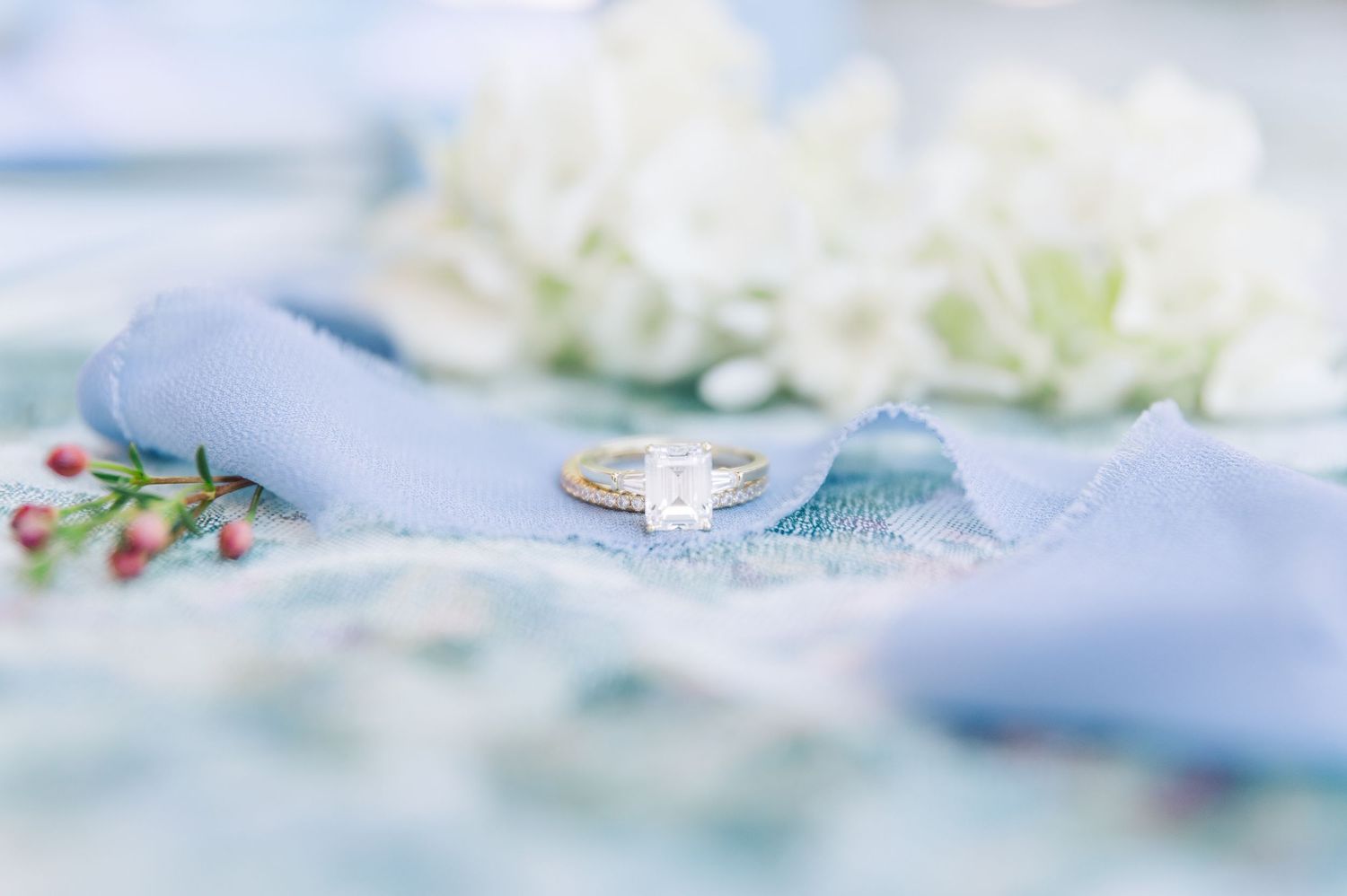 A delicate engagement ring rests on light blue fabric surrounded by soft white flowers and tiny red berries.