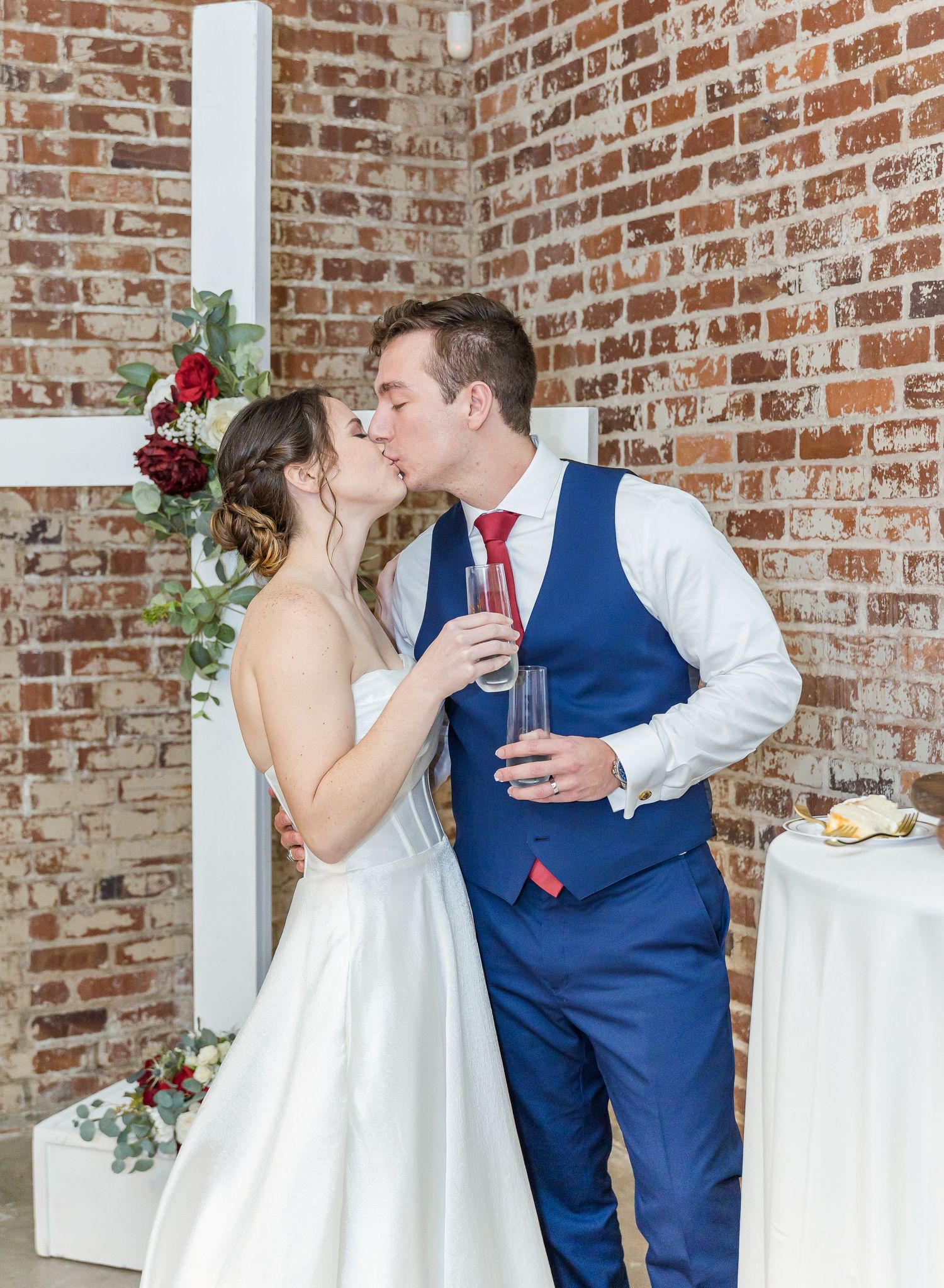 A wedding couple shares a kiss in front of an exposed brick wall decorated with red roses and greenery.