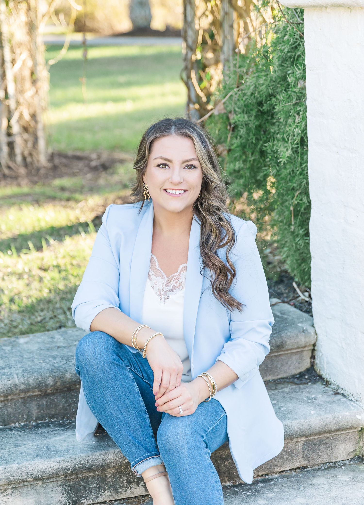 A person in a light blue blazer and jeans sits casually on outdoor stone steps next to greenery in natural lighting.