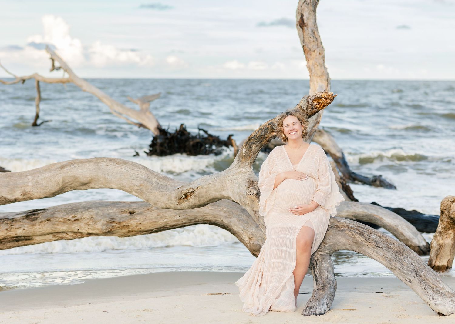 A pale pink maternity dress flows in the ocean breeze as someone poses on driftwood at a scenic beach.