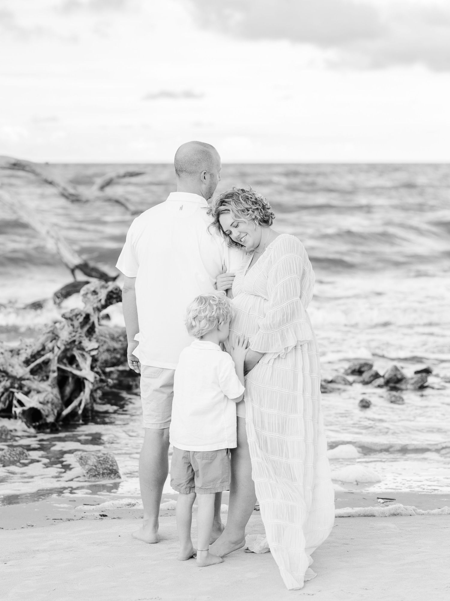 A seaside family portrait captures a tender moment along the water's edge at sunset.