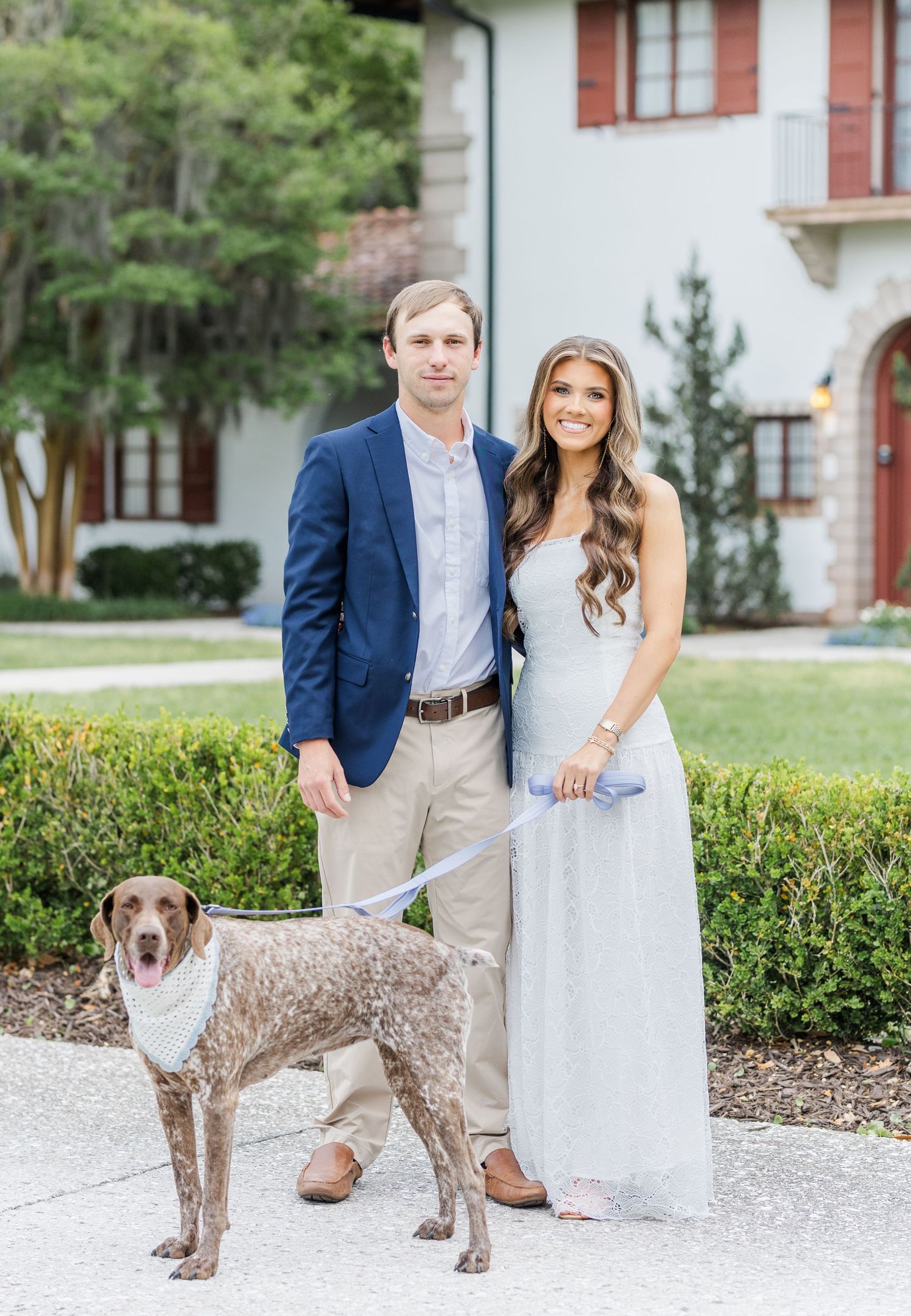 Couple in formal attire posing with spotted dog on leash in front of elegant white estate.