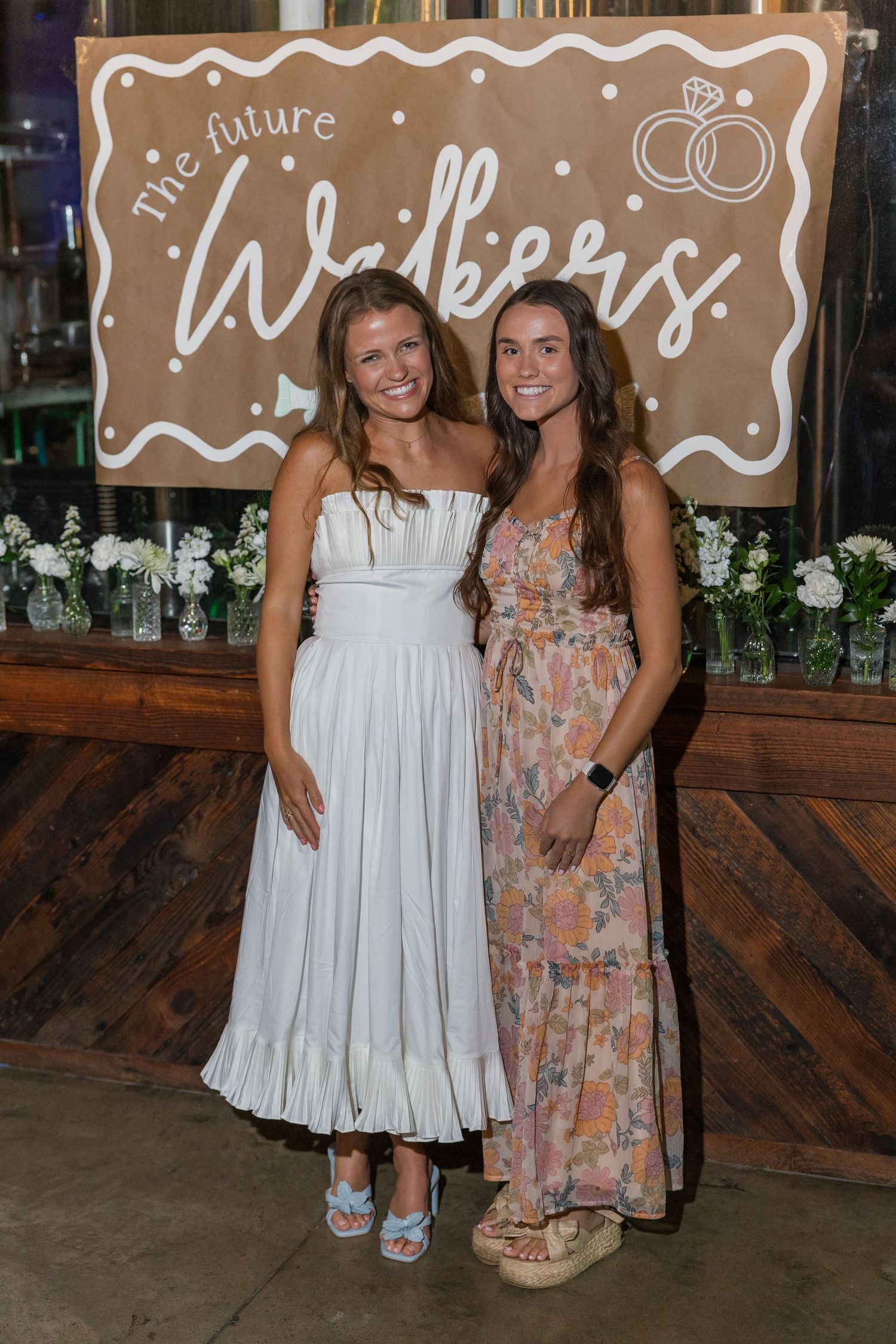 Two smiling women in white and floral dresses pose at The Future Walkers engagement celebration.