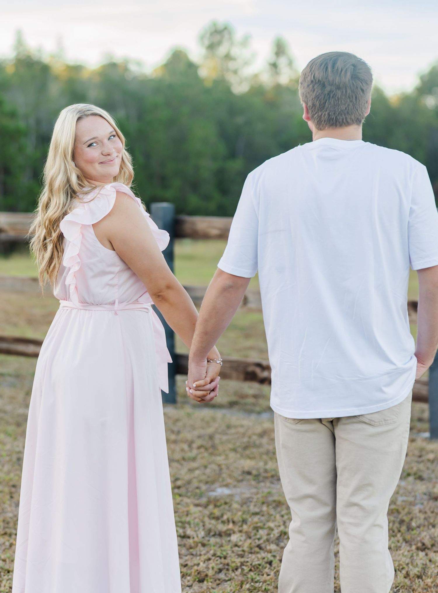 Couple in coordinating white and pink outfits walking hand in hand through a rustic outdoor setting.