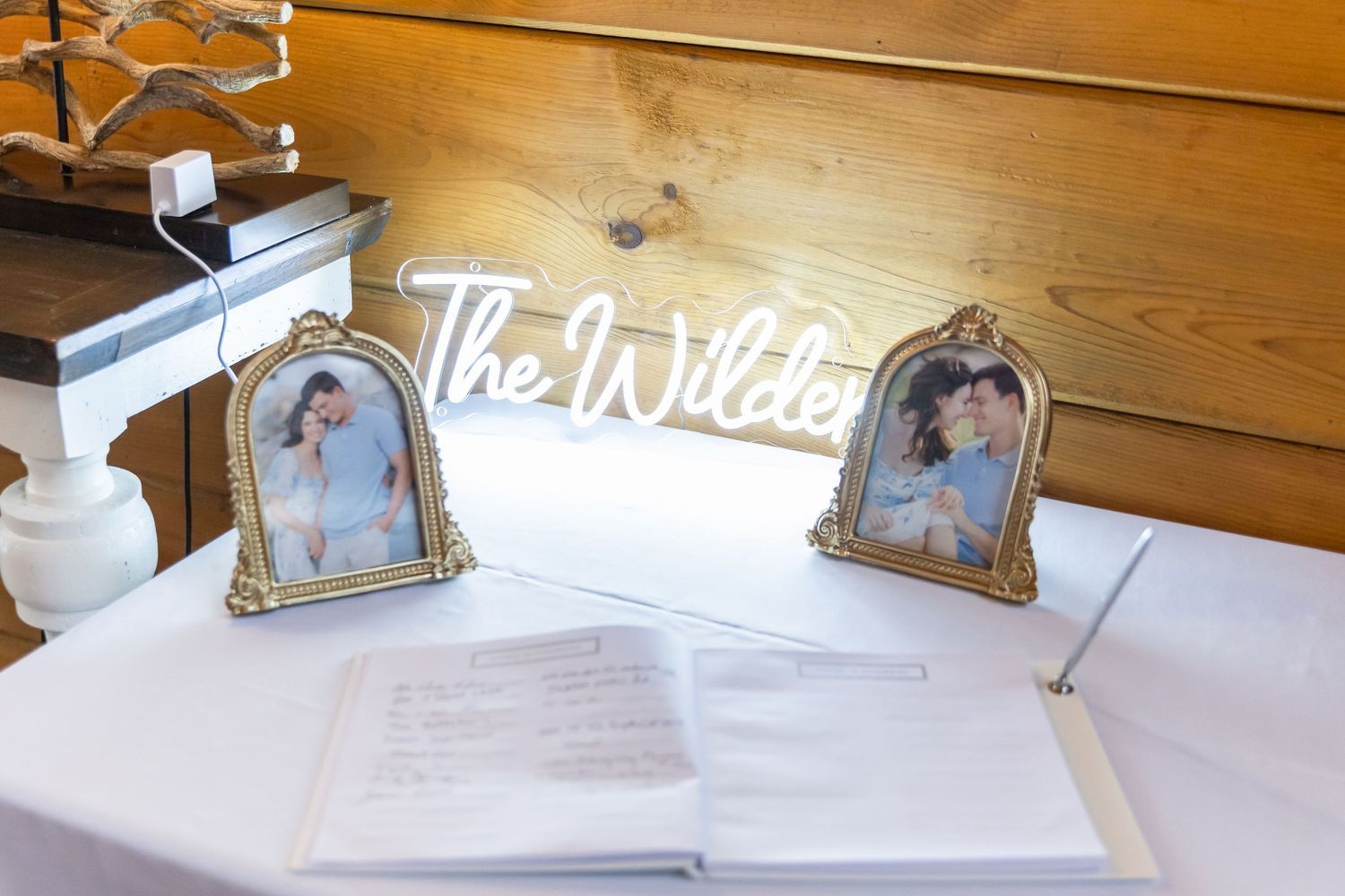 Wedding guest book table with neon 'The Wildes' sign and decorative frames on rustic wooden backdrop.