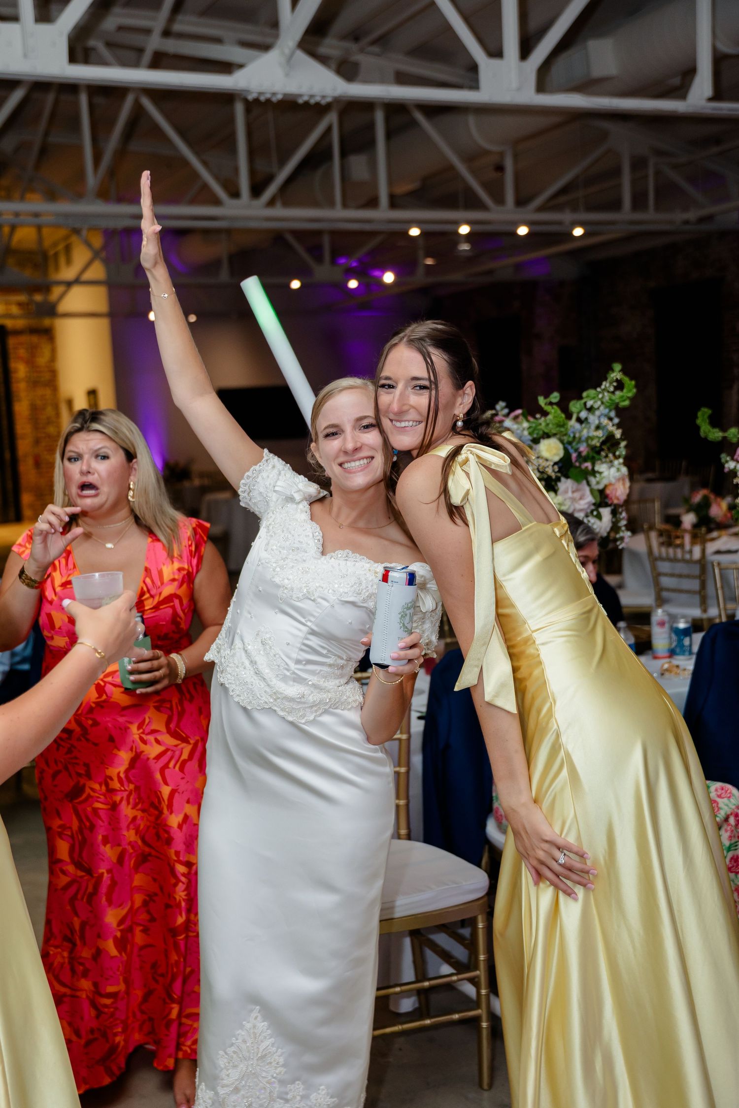 Wedding guests in red and yellow dresses dancing together at a reception in an industrial venue with exposed brick walls.