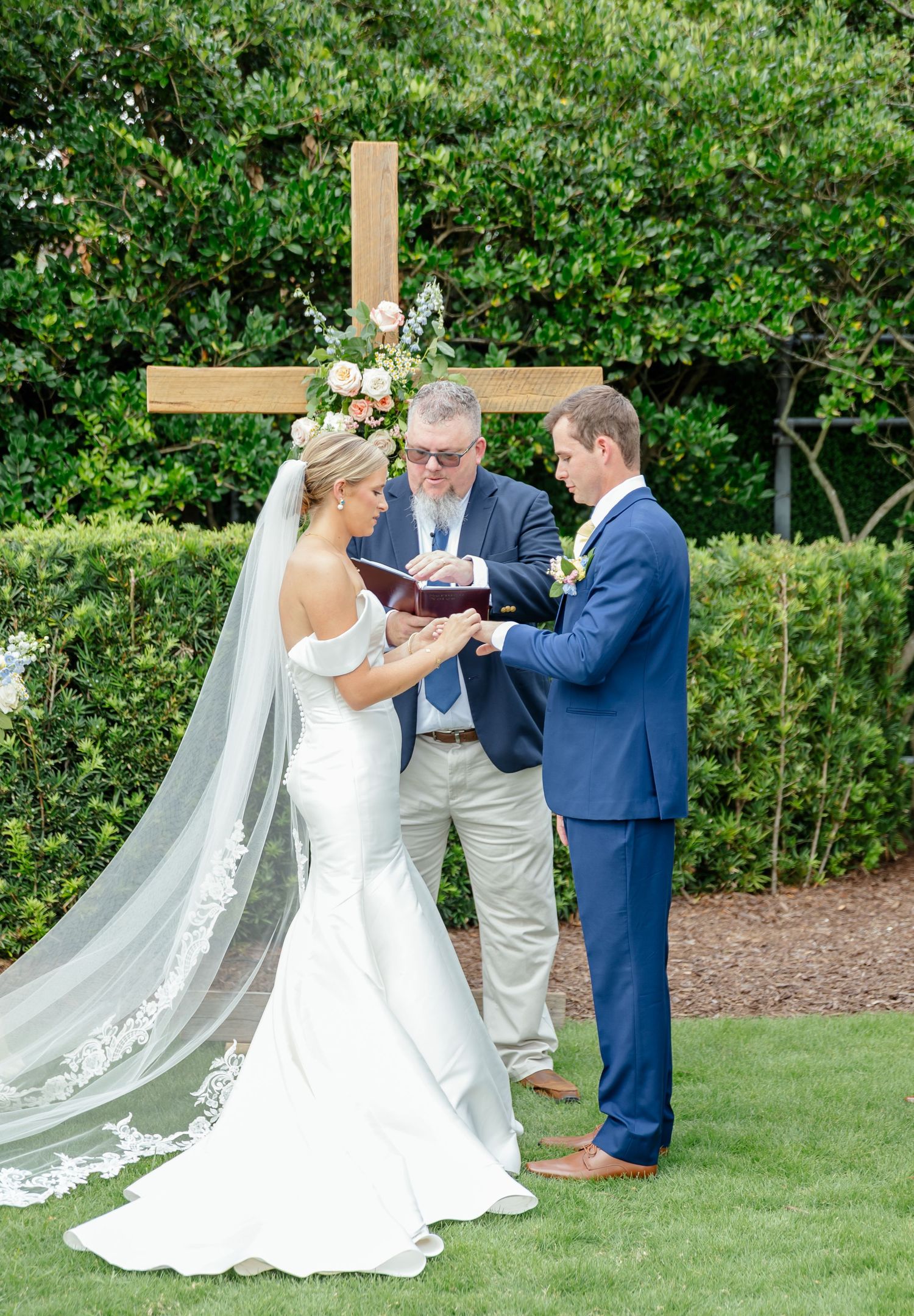 A wedding ceremony takes place outdoors on a lush green lawn with a beautiful white lace veil trailing behind the bride.