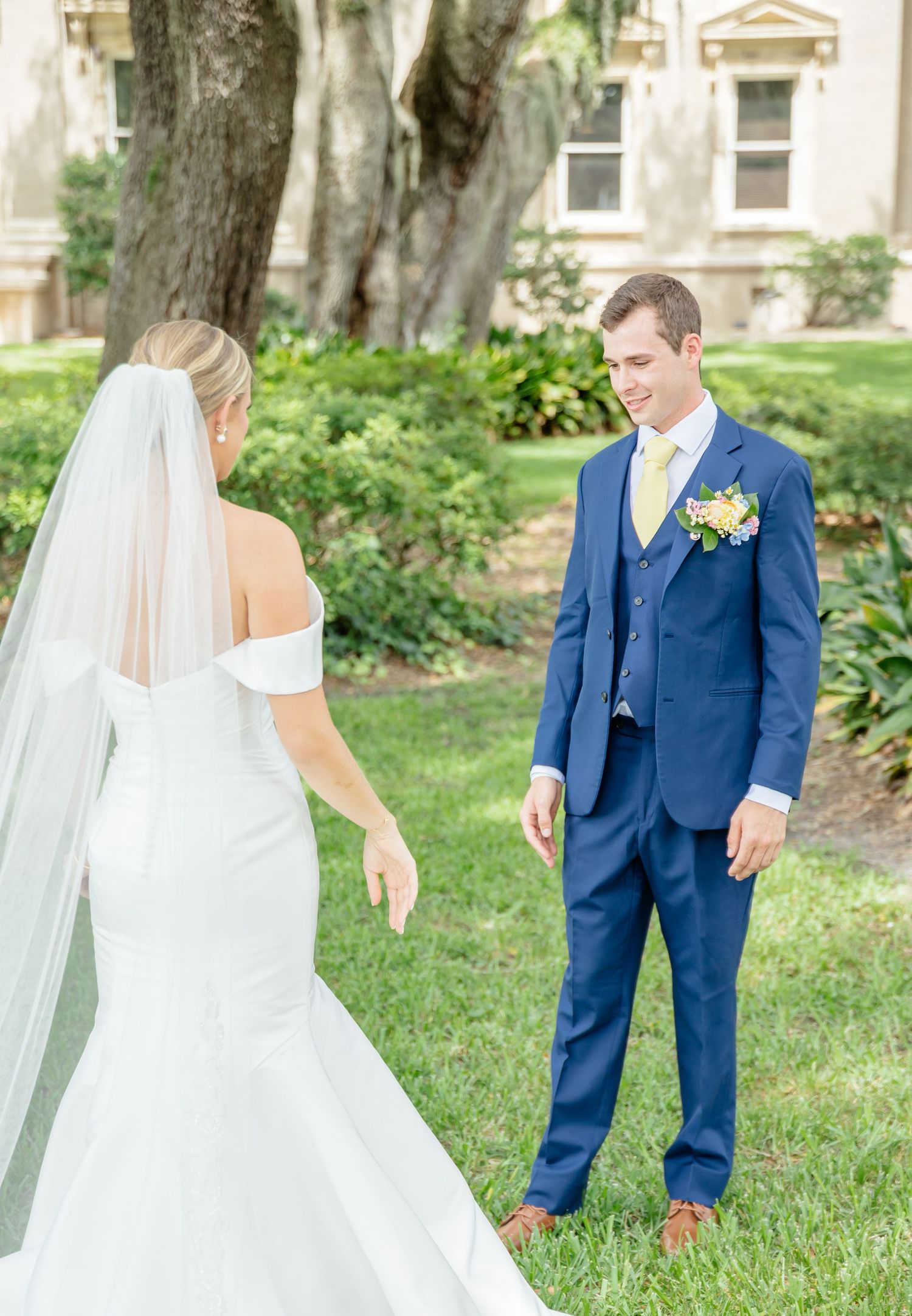 A first look moment between a couple in wedding attire on a grassy lawn with trees and buildings in the background.