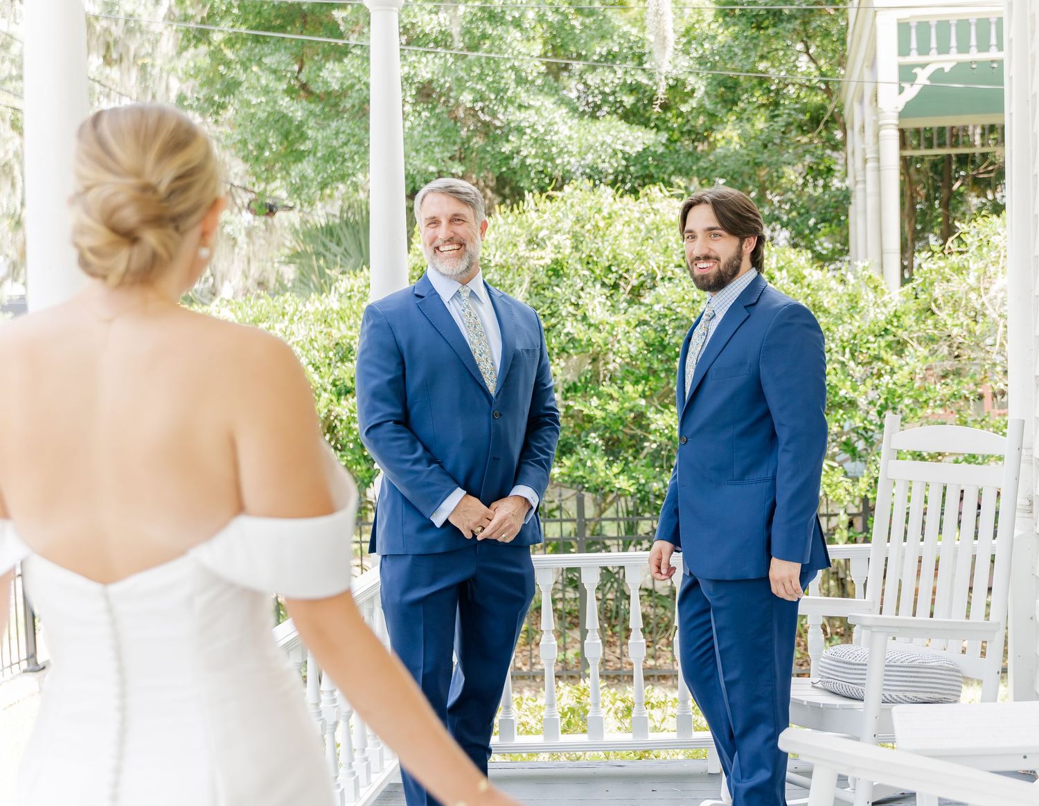 A wedding scene on a white porch with people in blue suits and a white wedding dress during a sunny day.