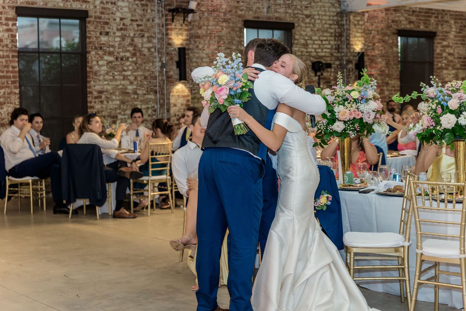 Wedding celebration in a rustic brick venue with floral arrangements and gold chairs.