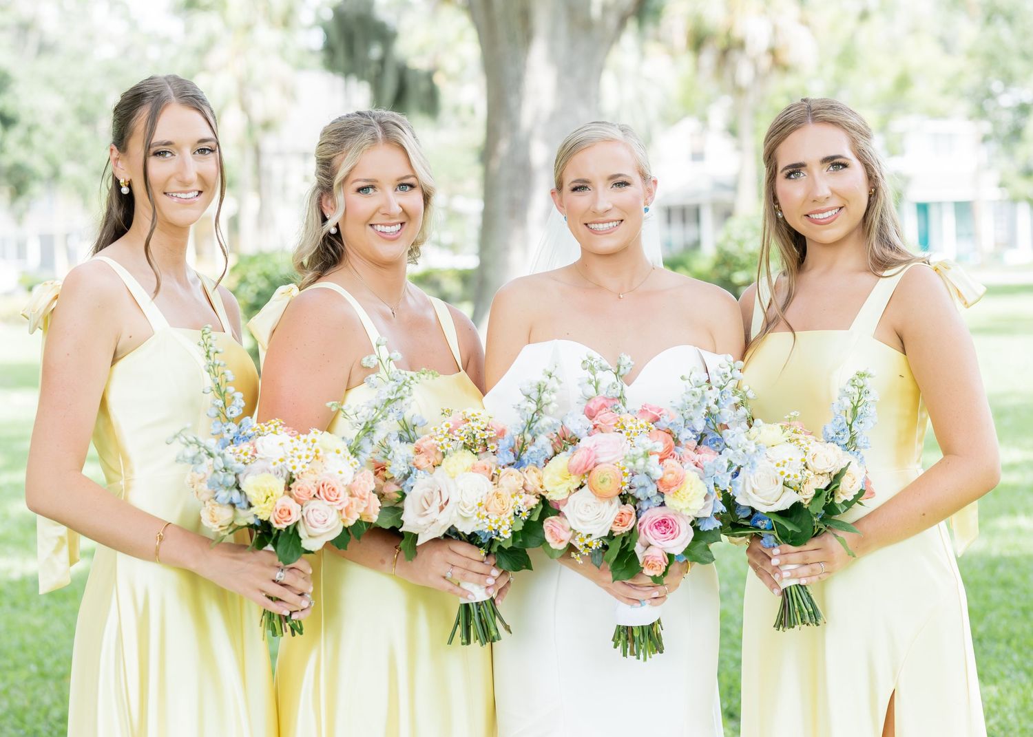 Bridesmaids in pale yellow dresses hold pastel bouquets while posing with bride at outdoor garden wedding.