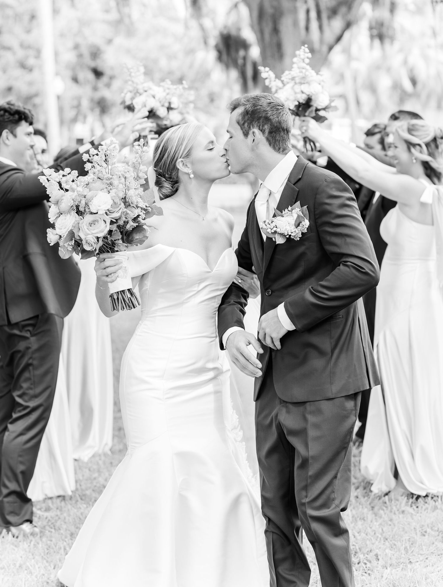 Black and white photo of a romantic wedding kiss moment with bridal party in background at an outdoor ceremony.