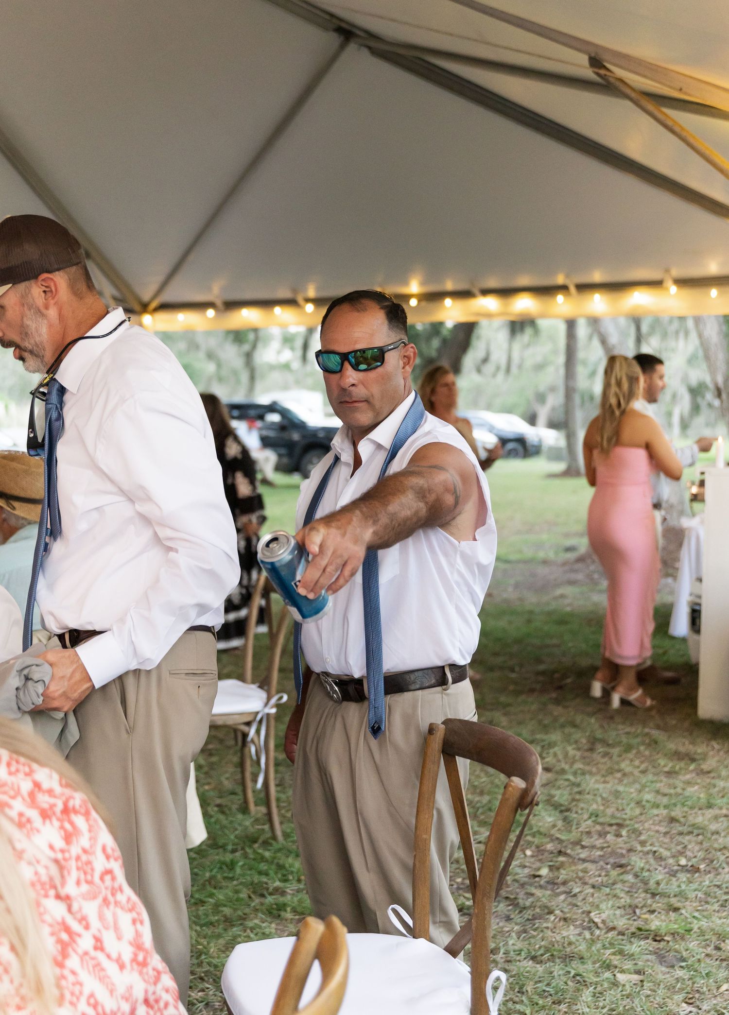 People gather under an outdoor event tent with string lights, dressed in casual summer attire.