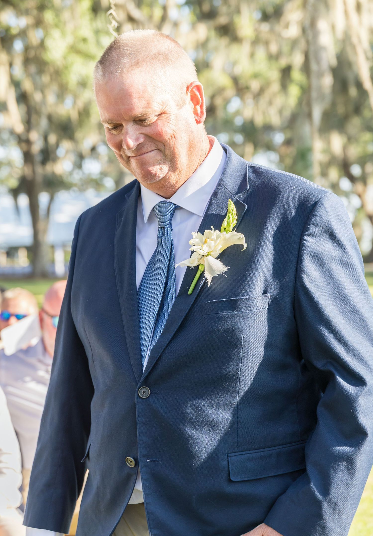 A person wearing a navy suit with a light blue tie and boutonniere smiles during an outdoor wedding ceremony.