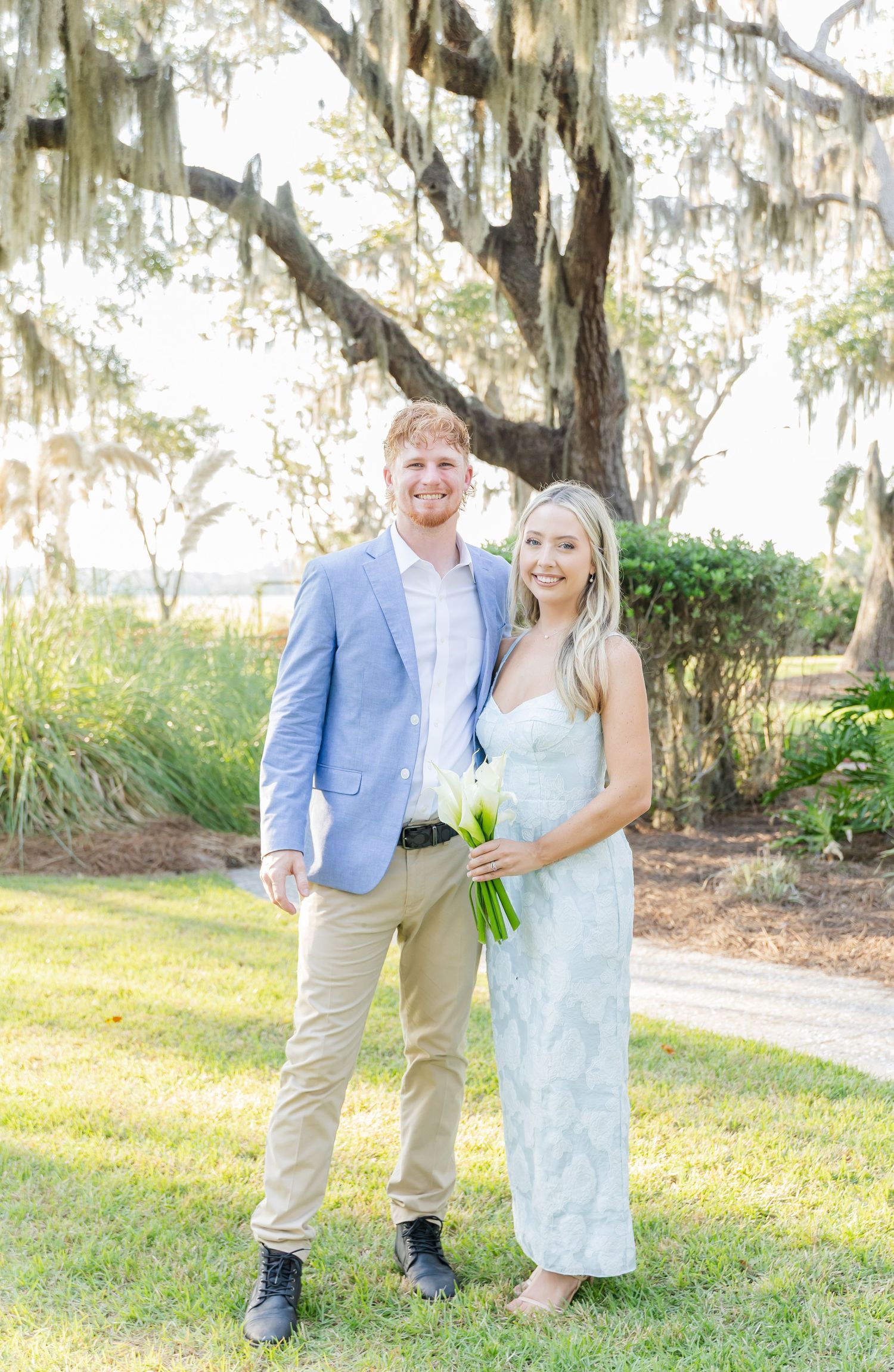 A couple poses outdoors beneath Spanish moss-draped oak trees in a romantic garden setting.