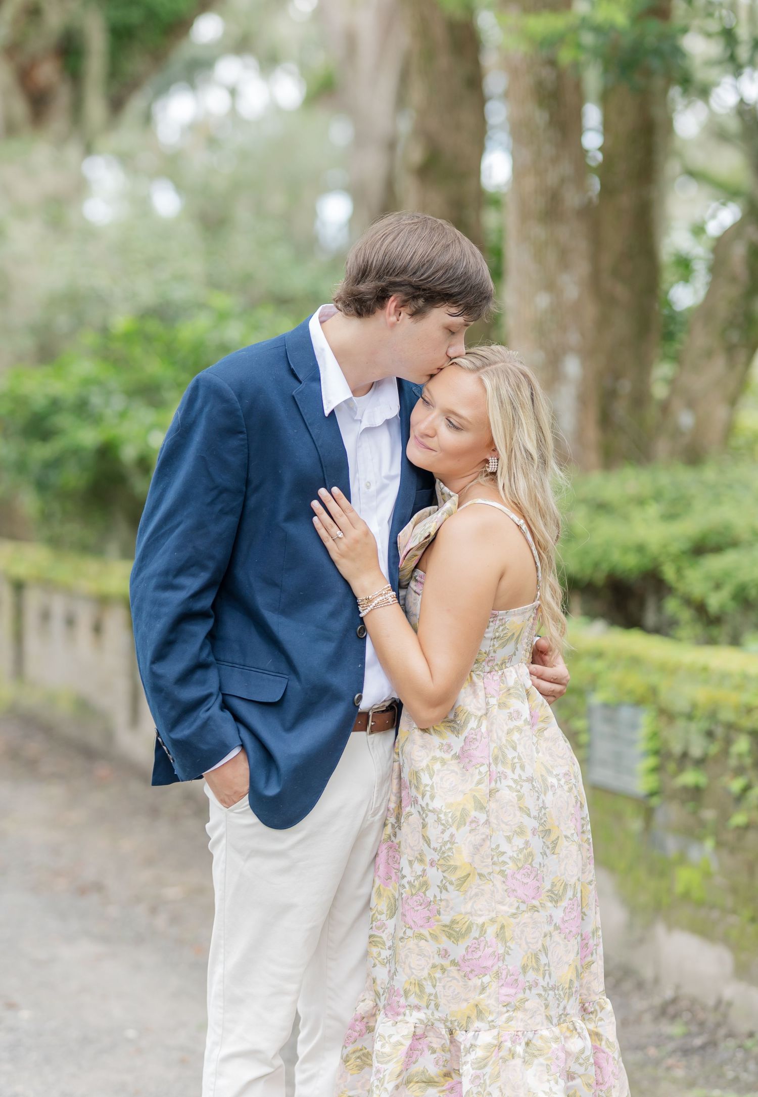 A romantic couple embraces on a tree-lined path, with the person in a navy blazer and white pants and their partner in a floral dress.
