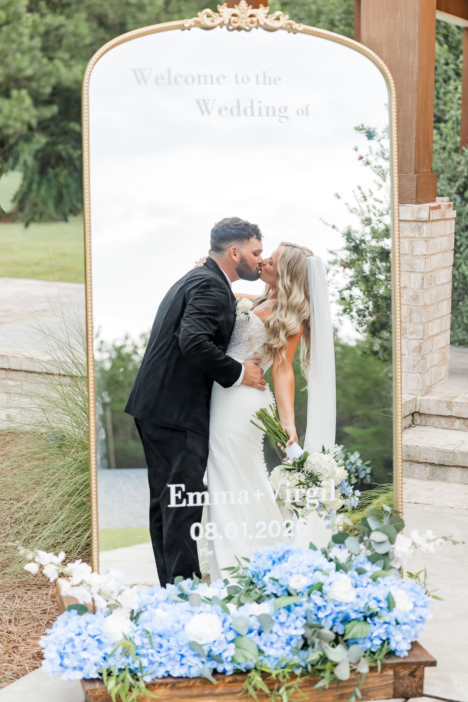 Bride and groom kissing in front of gold welcome mirror with blue hydrangea floral arrangements below.