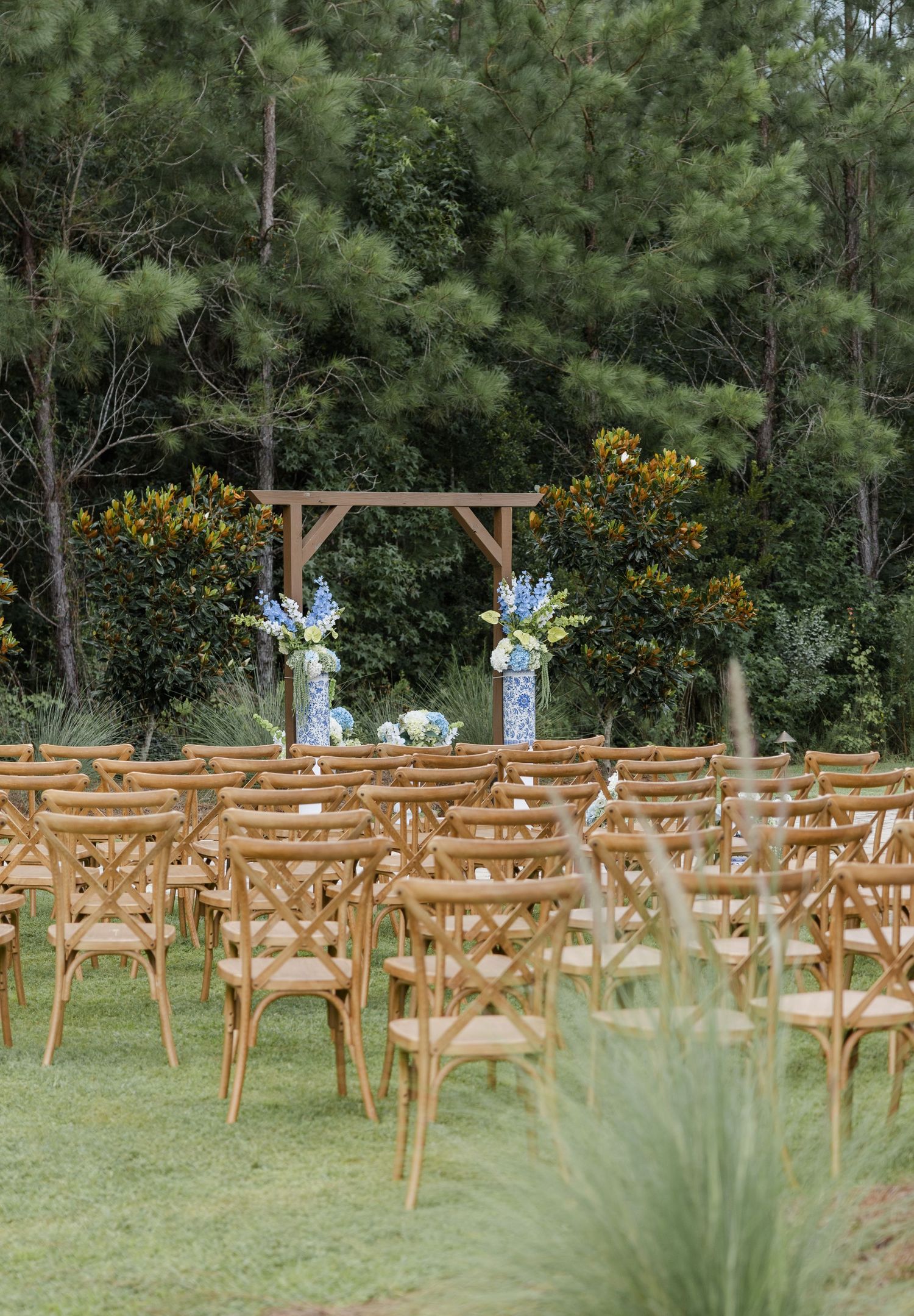 Outdoor wedding ceremony setup with wooden cross-back chairs, wooden arch, and blue floral arrangements against a lush green forest backdrop