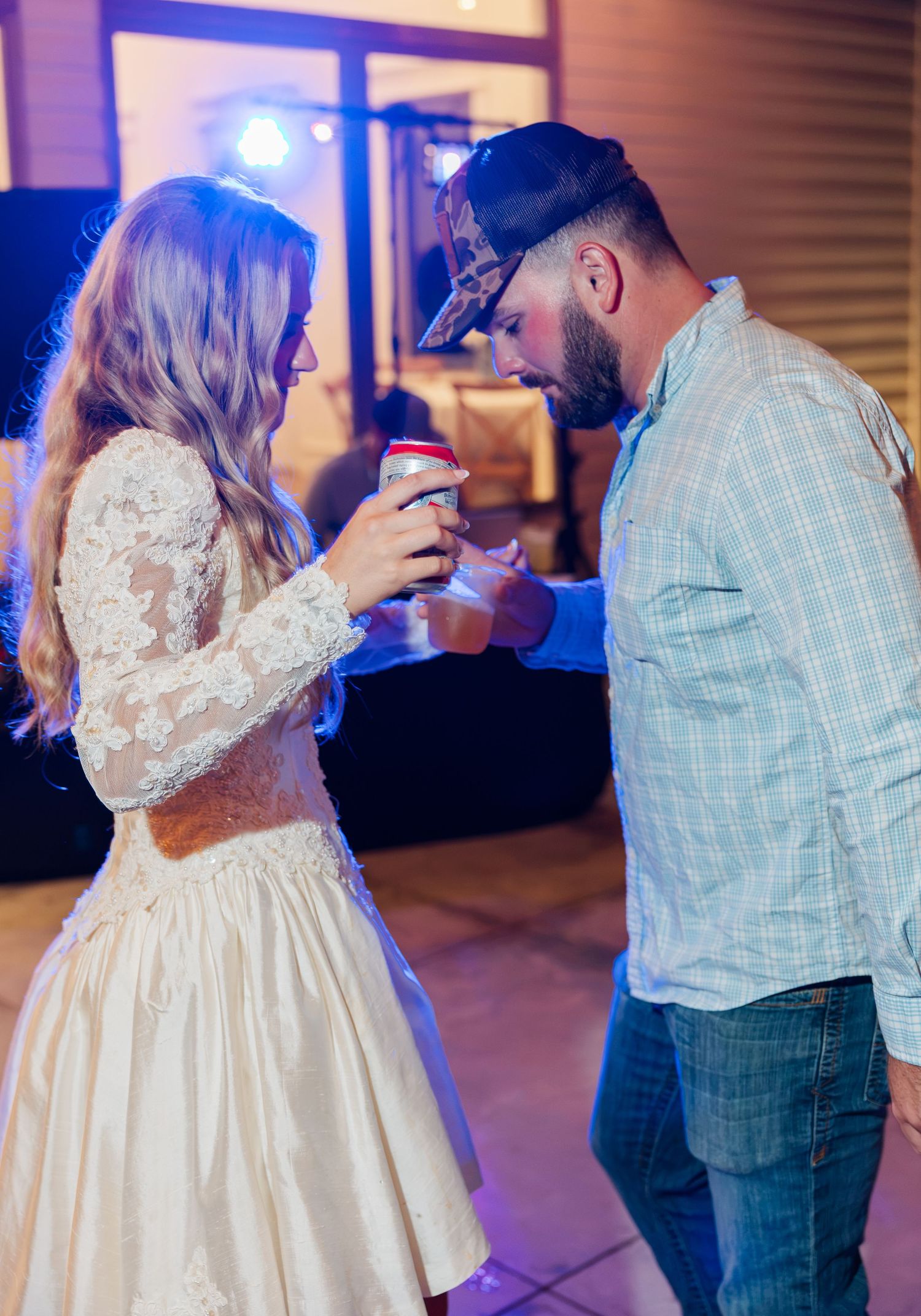 Bride in lace dress and man in plaid shirt share a close dance moment at lively wedding reception.